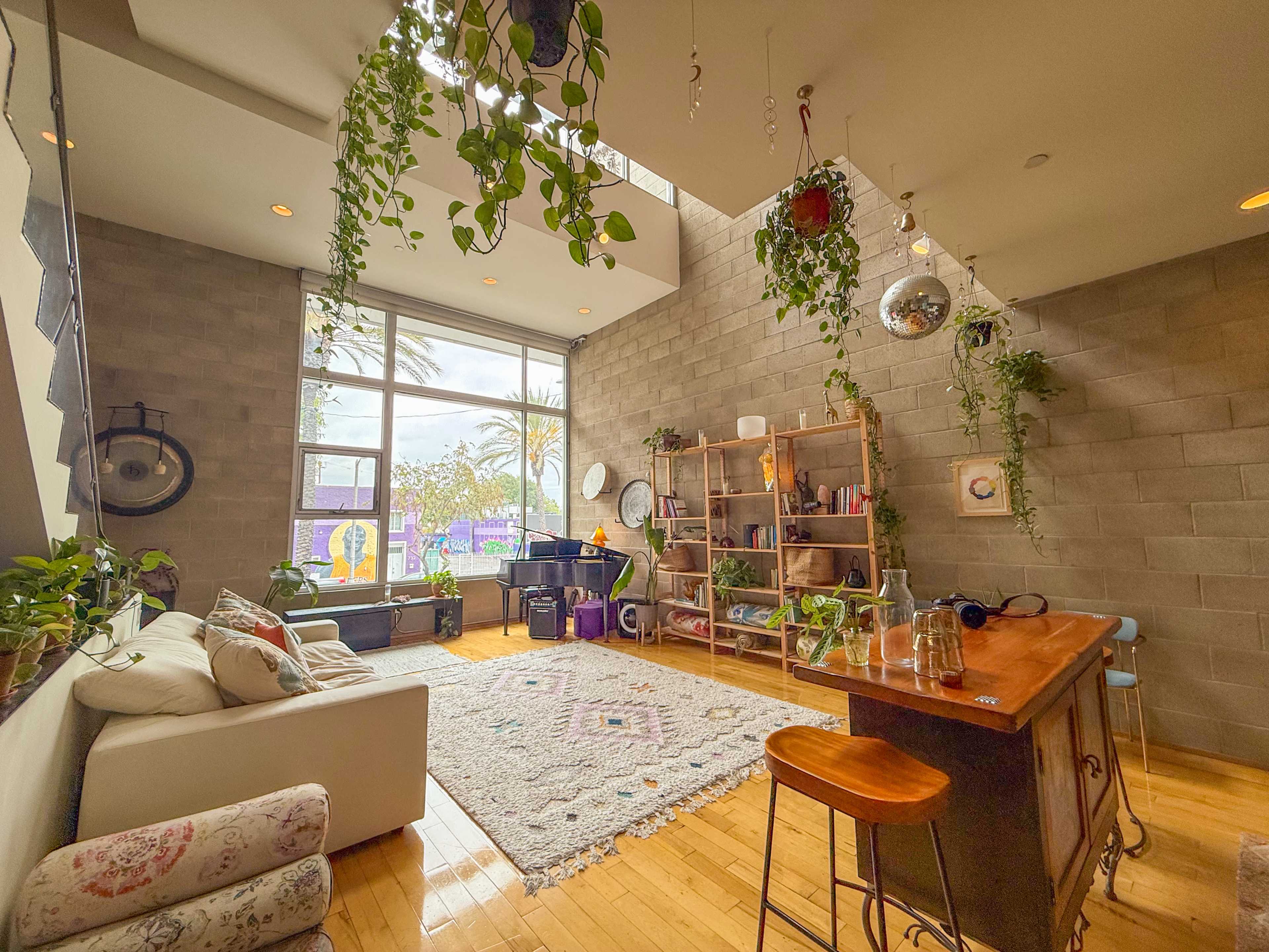 The image shows a modern living space with a wooden dining table, a sofa, indoor plants, and large windows allowing natural light to illuminate the room.