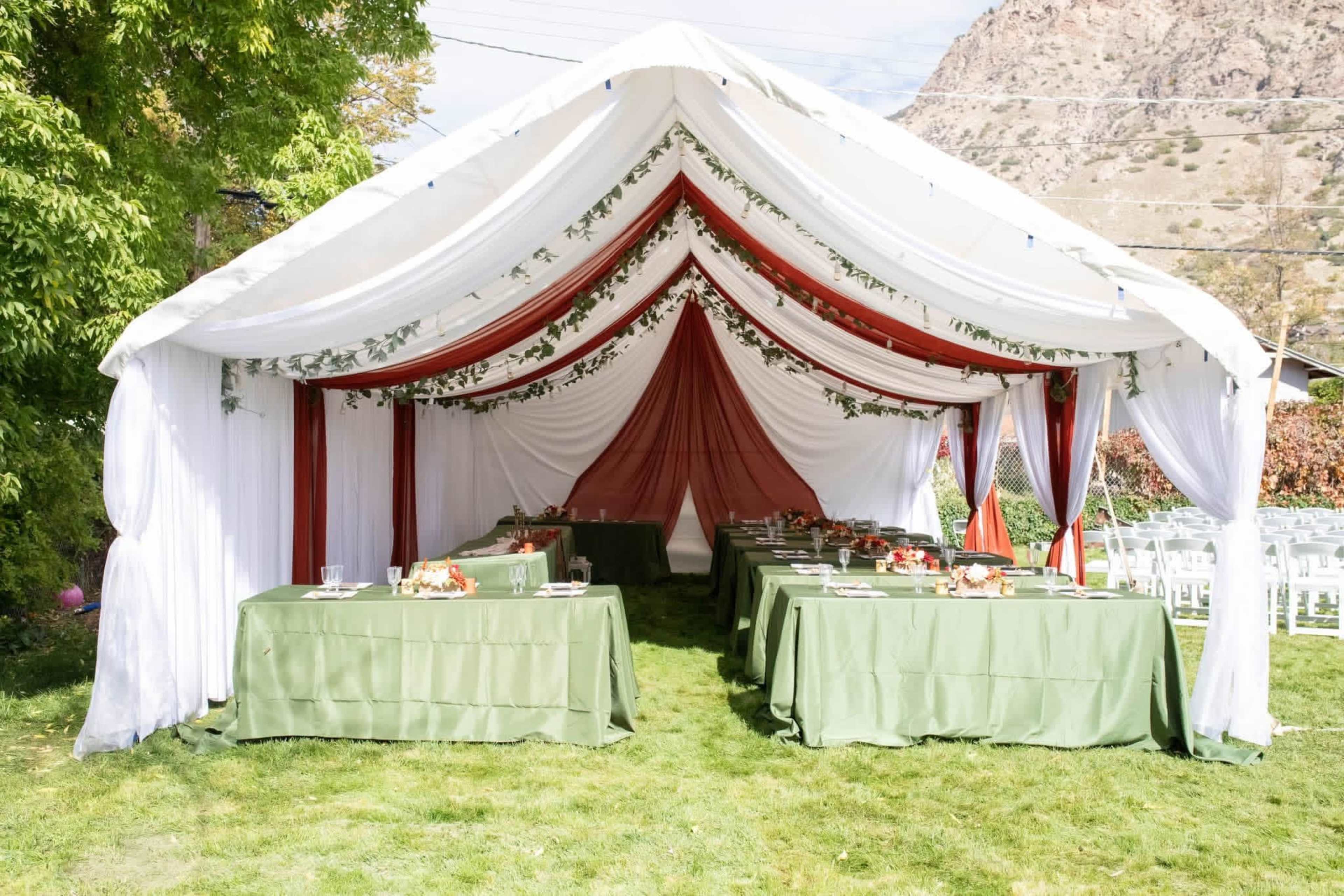 A white and red tent featuring green tables is set up on grass for a dining arrangement, surrounded by a mountainous landscape.