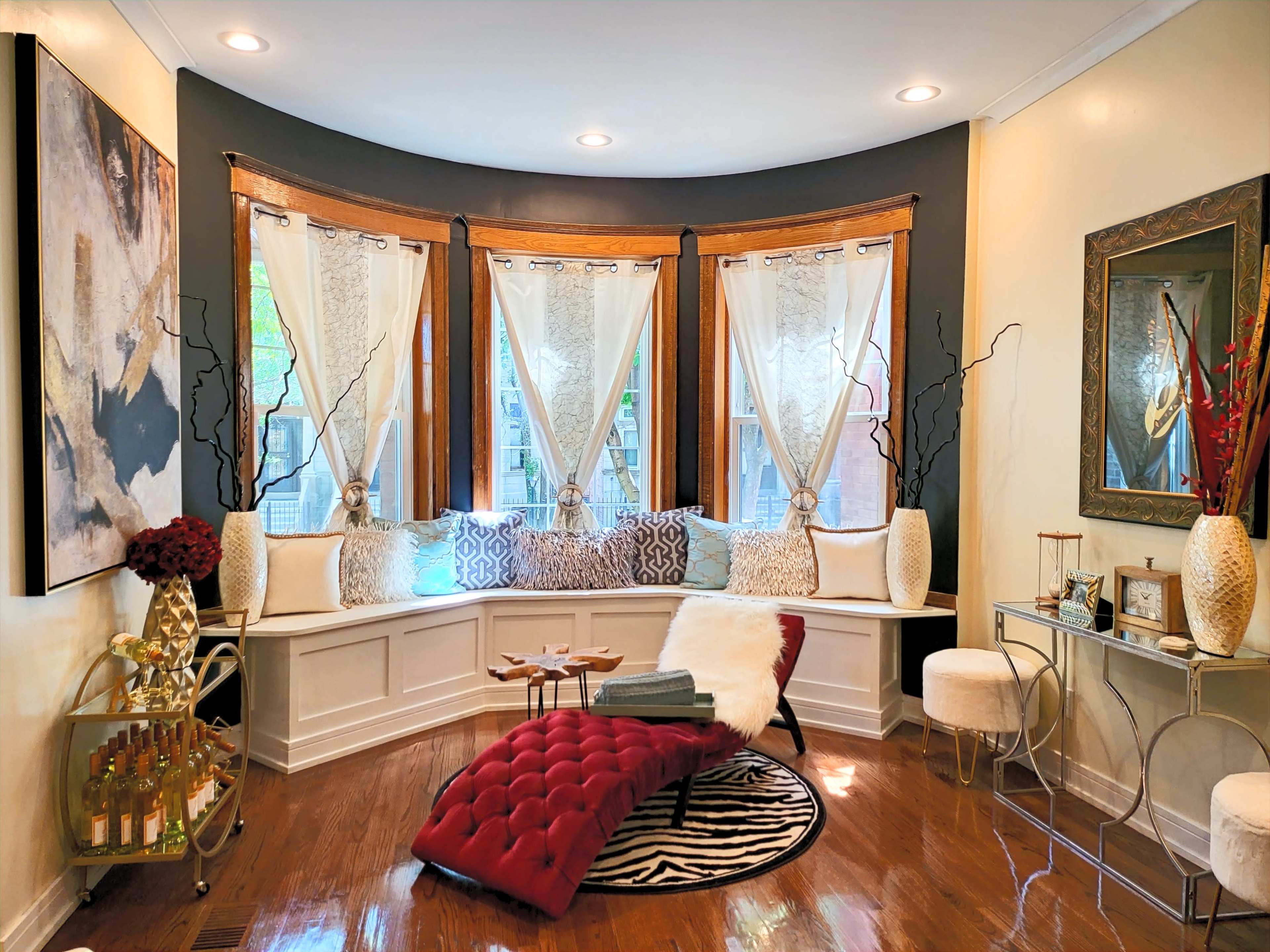The scene features a cozy nook with a curved window seat adorned with pillows, a fur throw, and a small table, adjacent to a decorative mirror and a stylish side table.