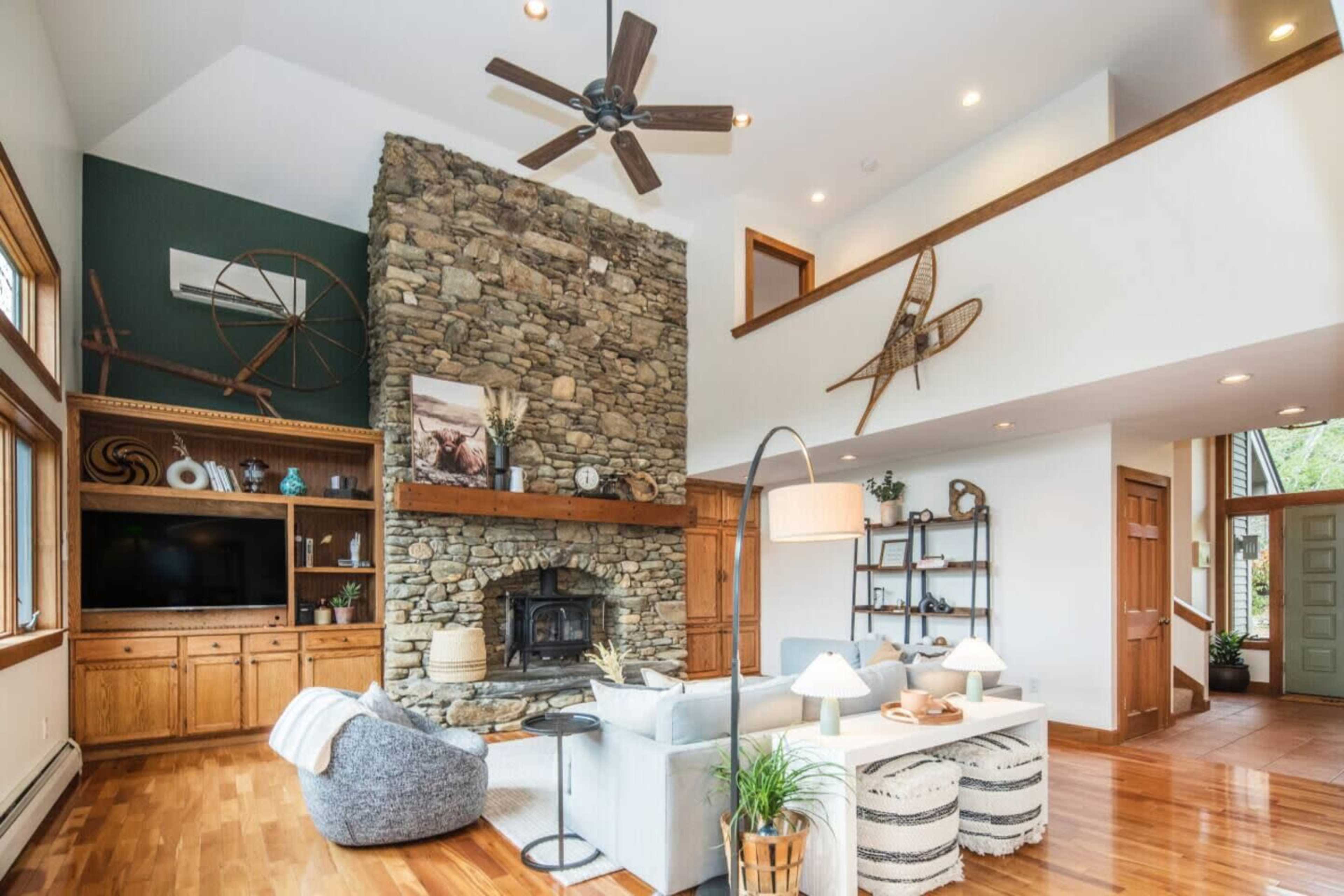The image shows a modern living room featuring a stone fireplace, a vaulted ceiling, wooden accents, and a mix of comfortable furniture arrangements.