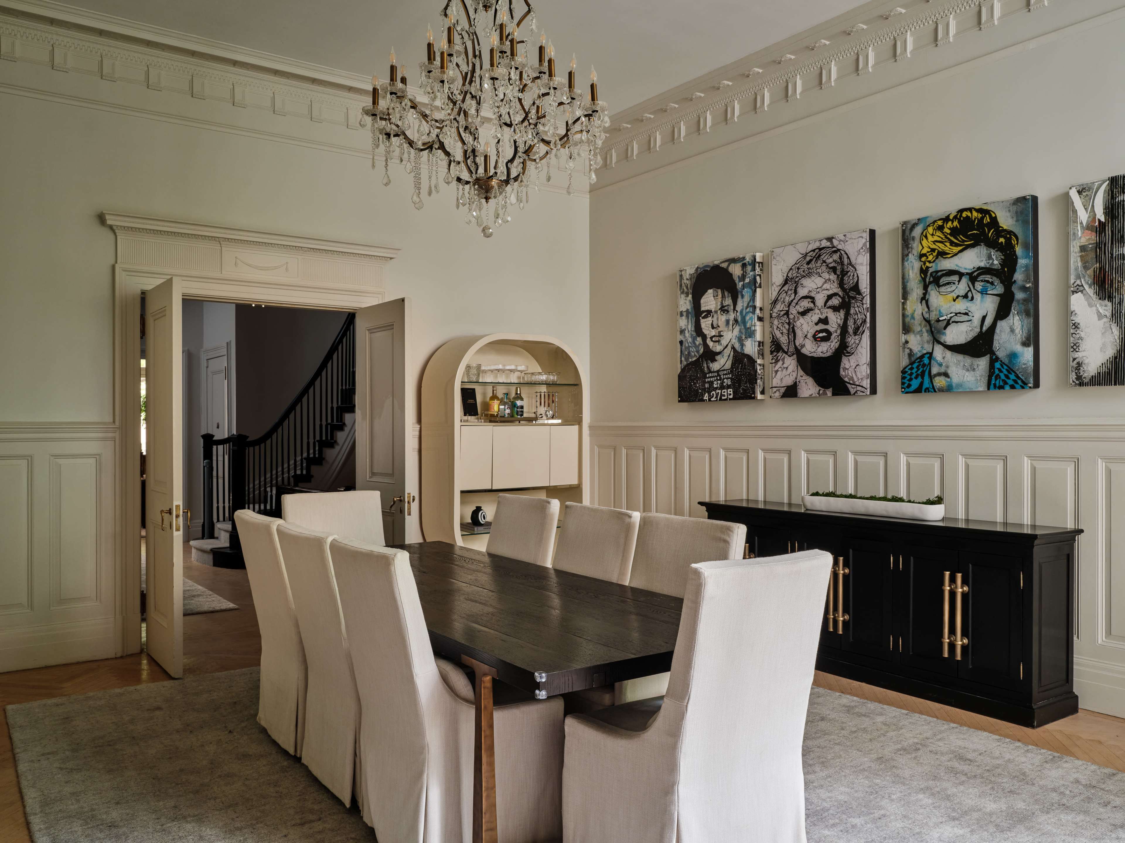 A dining room features a long wooden table surrounded by upholstered chairs, with a decorative chandelier overhead and art displayed on the walls.