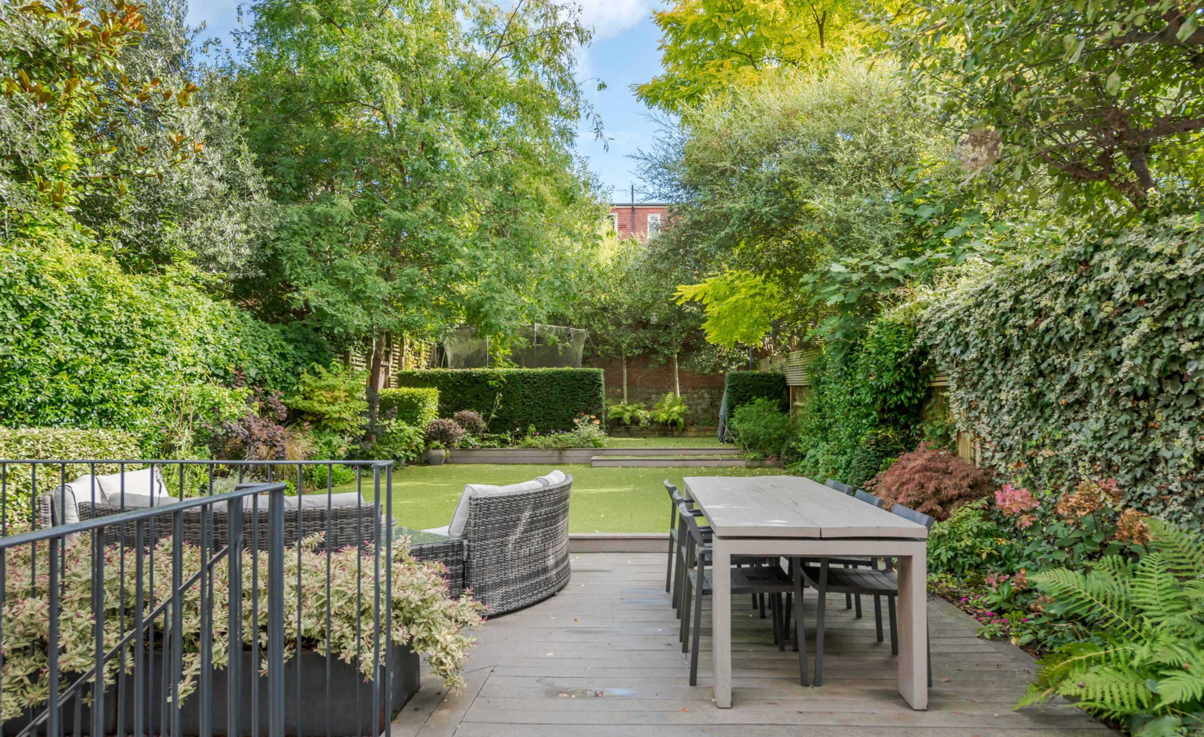 A well-maintained garden features a dining table and chairs on a deck, surrounded by lush greenery and neatly trimmed hedges.