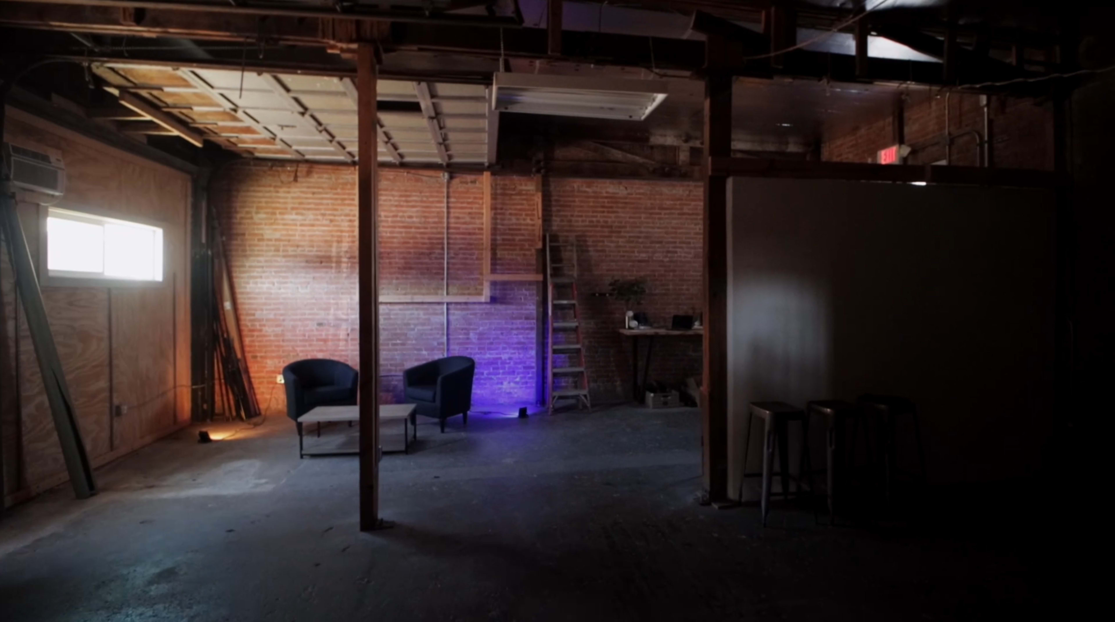 An empty loft space with exposed brick walls, two chairs, a small table, and a ladder against the wall.