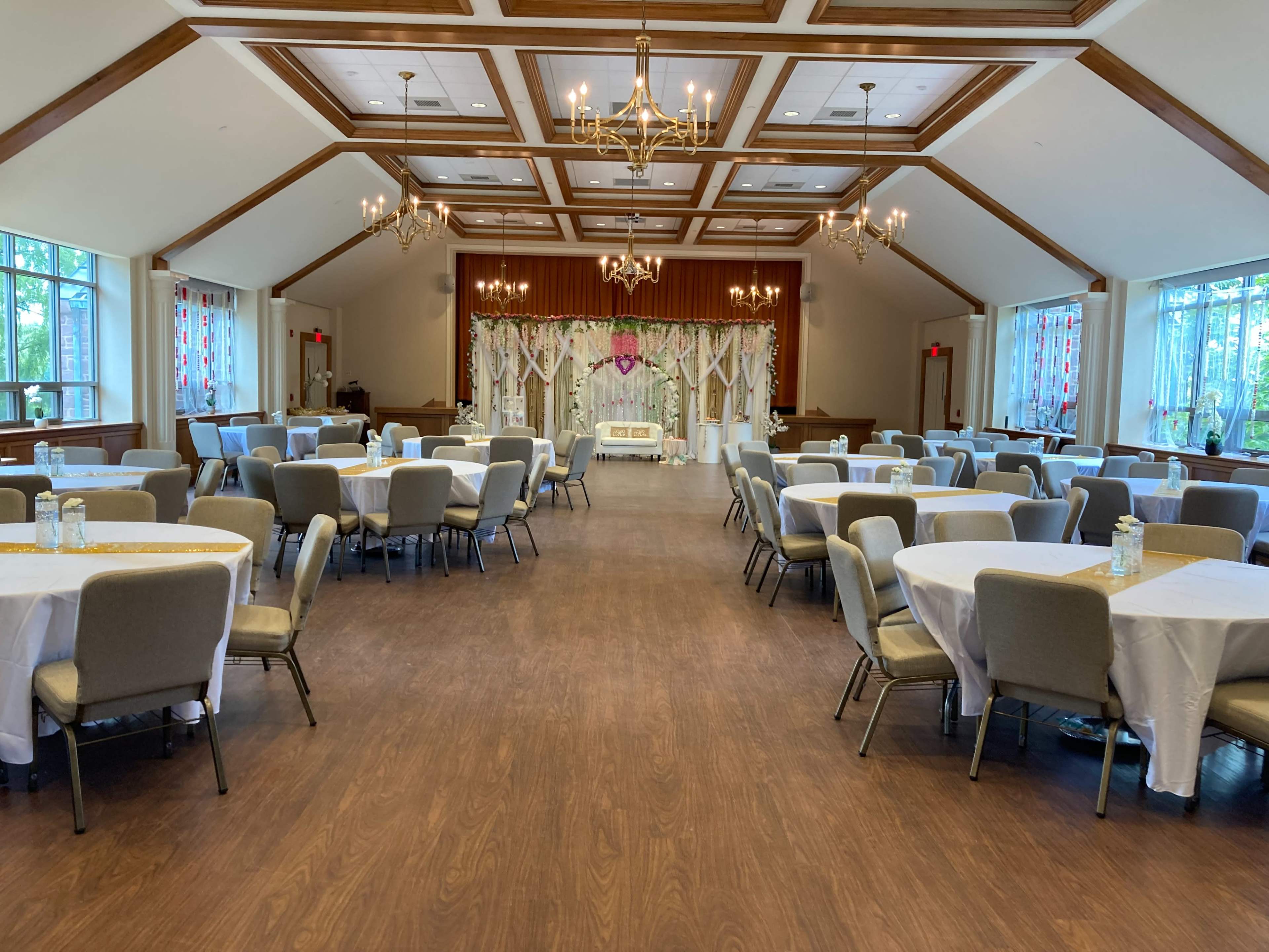 The image shows a brightly lit event hall with tables set for a gathering, featuring white tablecloths and gray chairs arranged around the space, and a decorated stage at the front.