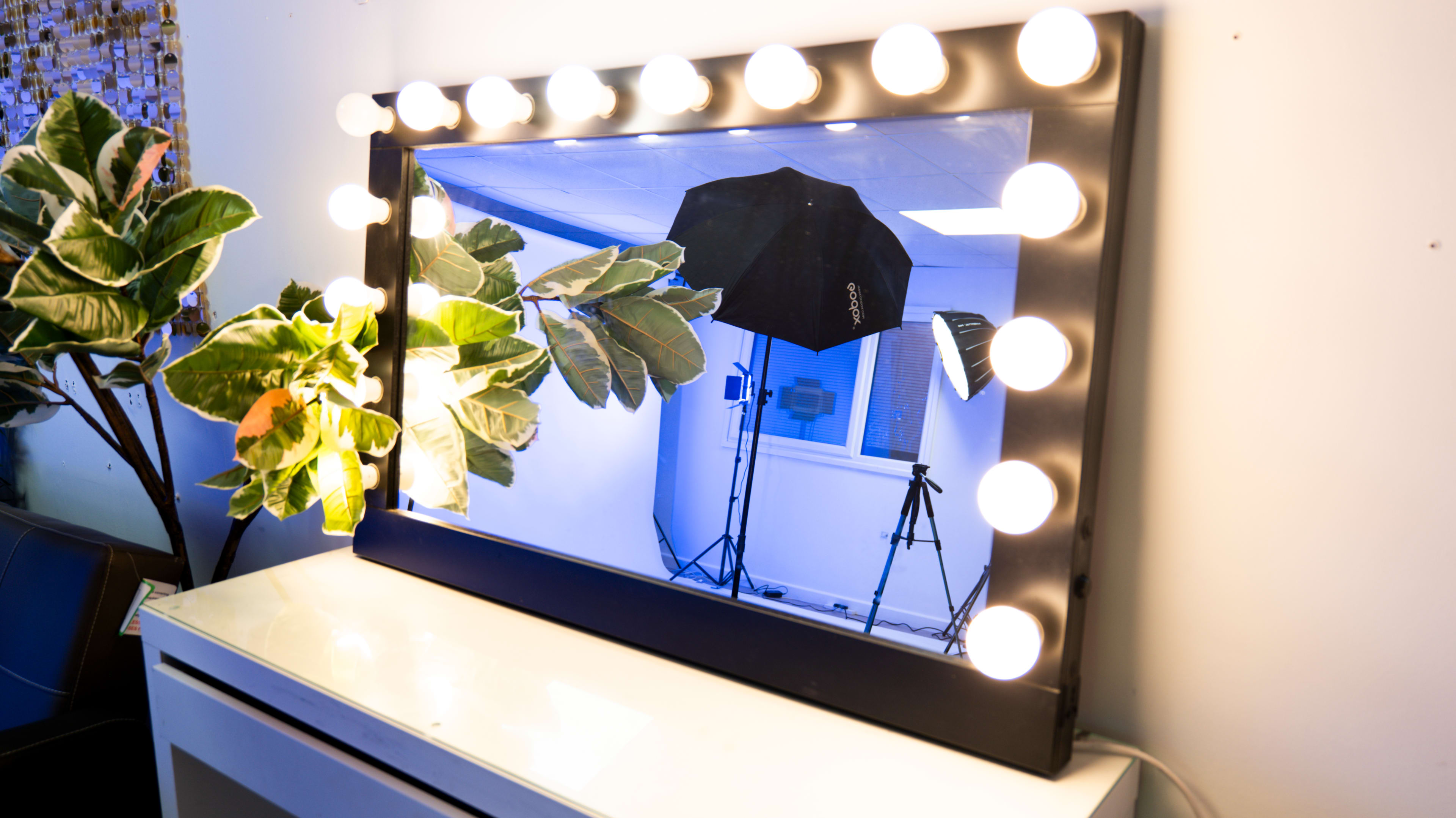 A large illuminated mirror stands on a white surface, reflecting a backdrop with a photography umbrella and tripods in a well-lit room.