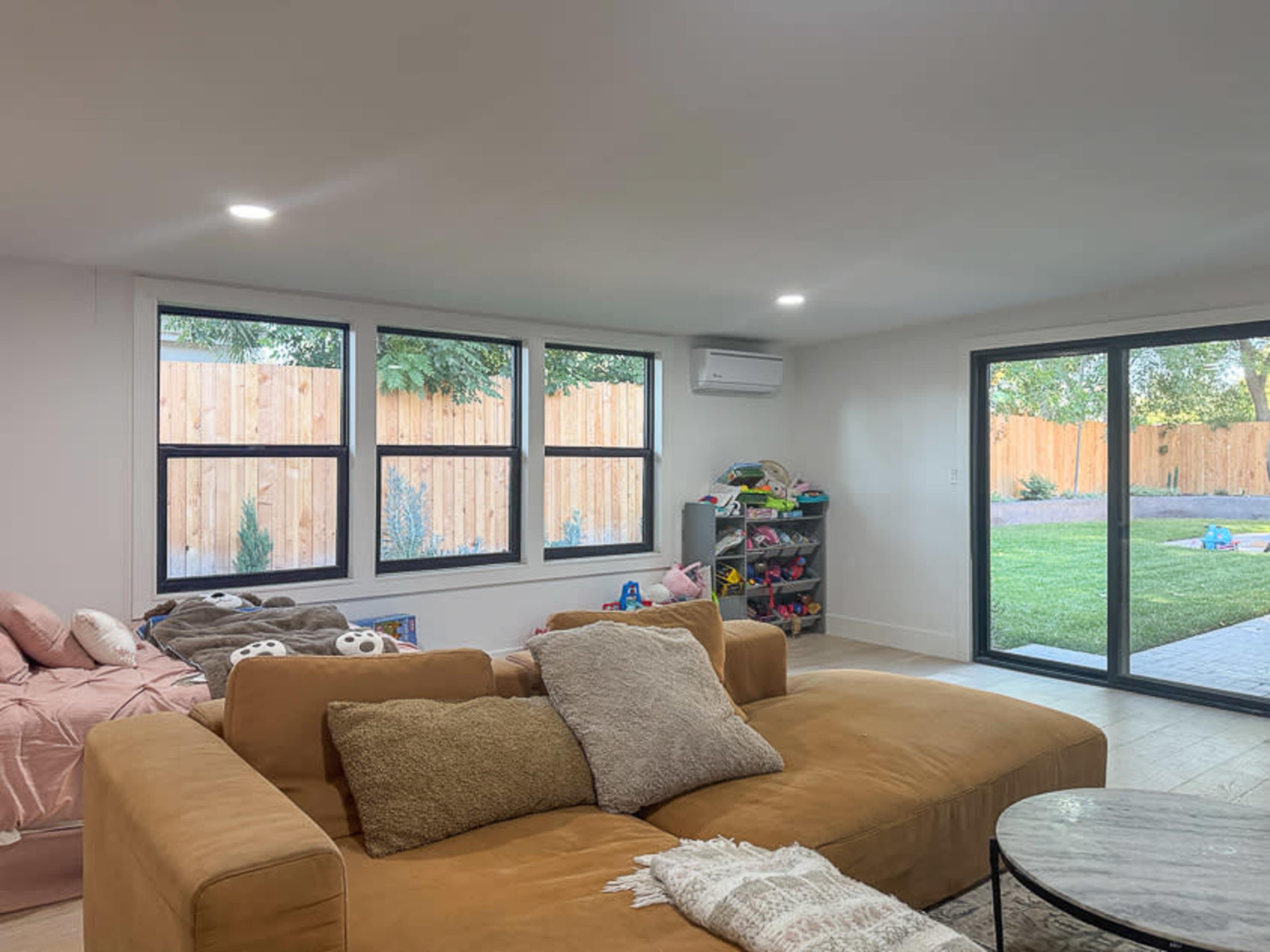 The image shows a modern living room with a large brown couch, a coffee table, and large windows overlooking a backyard with a wooden fence.