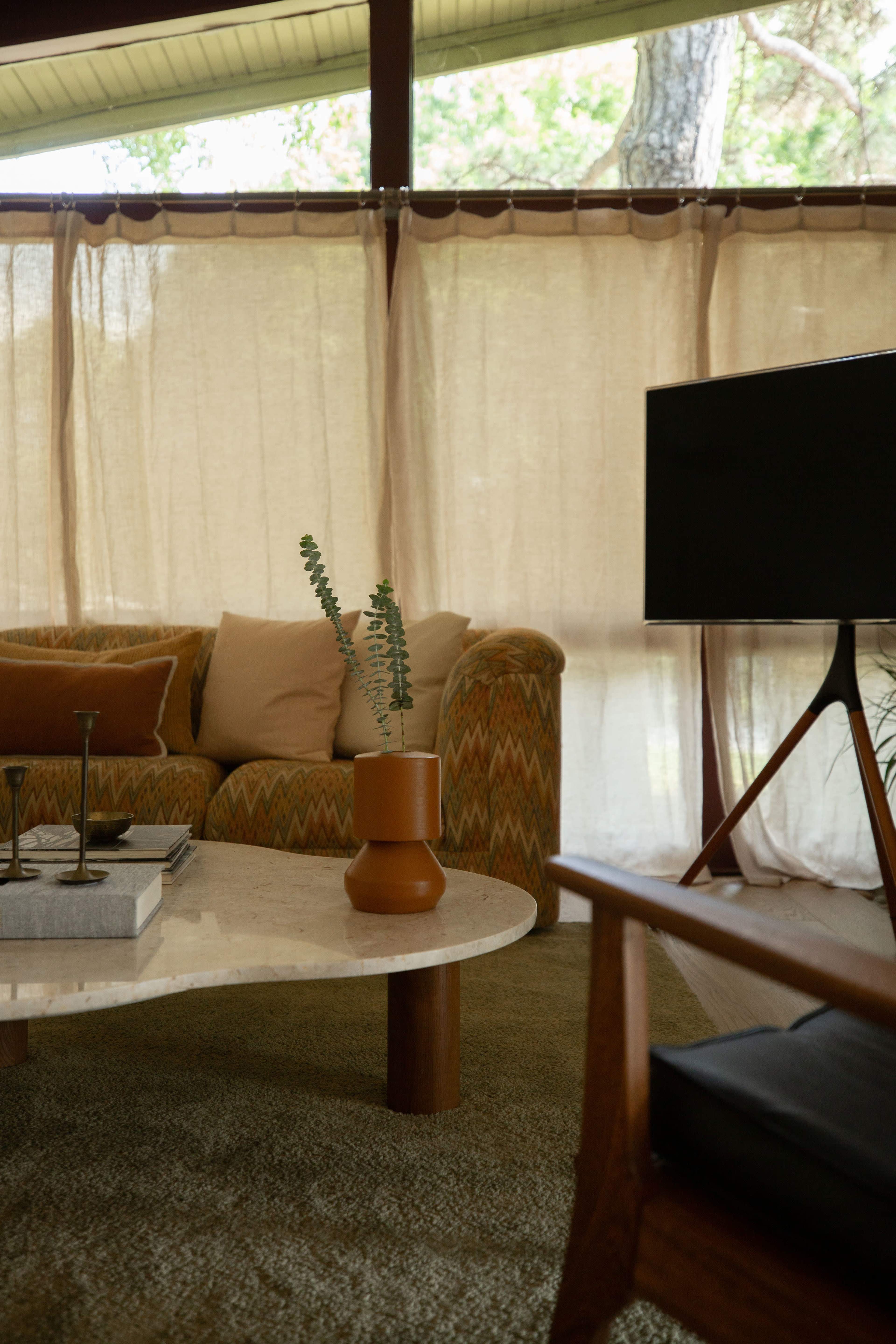 A cozy seating area features a patterned couch, a round marble coffee table, and a television beside sheer curtains filtering natural light.