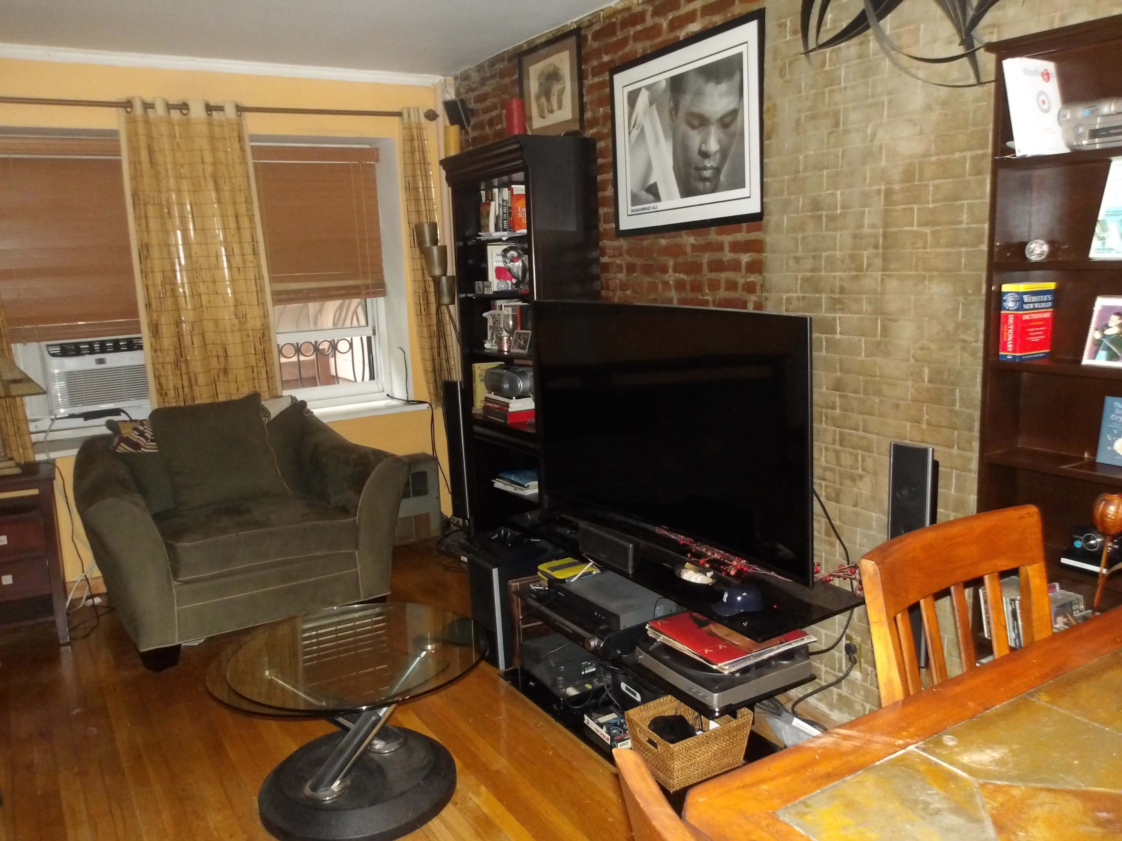 A small living room featuring a green armchair, a large television on a black entertainment unit, a glass coffee table, and wooden furniture against a brick wall.