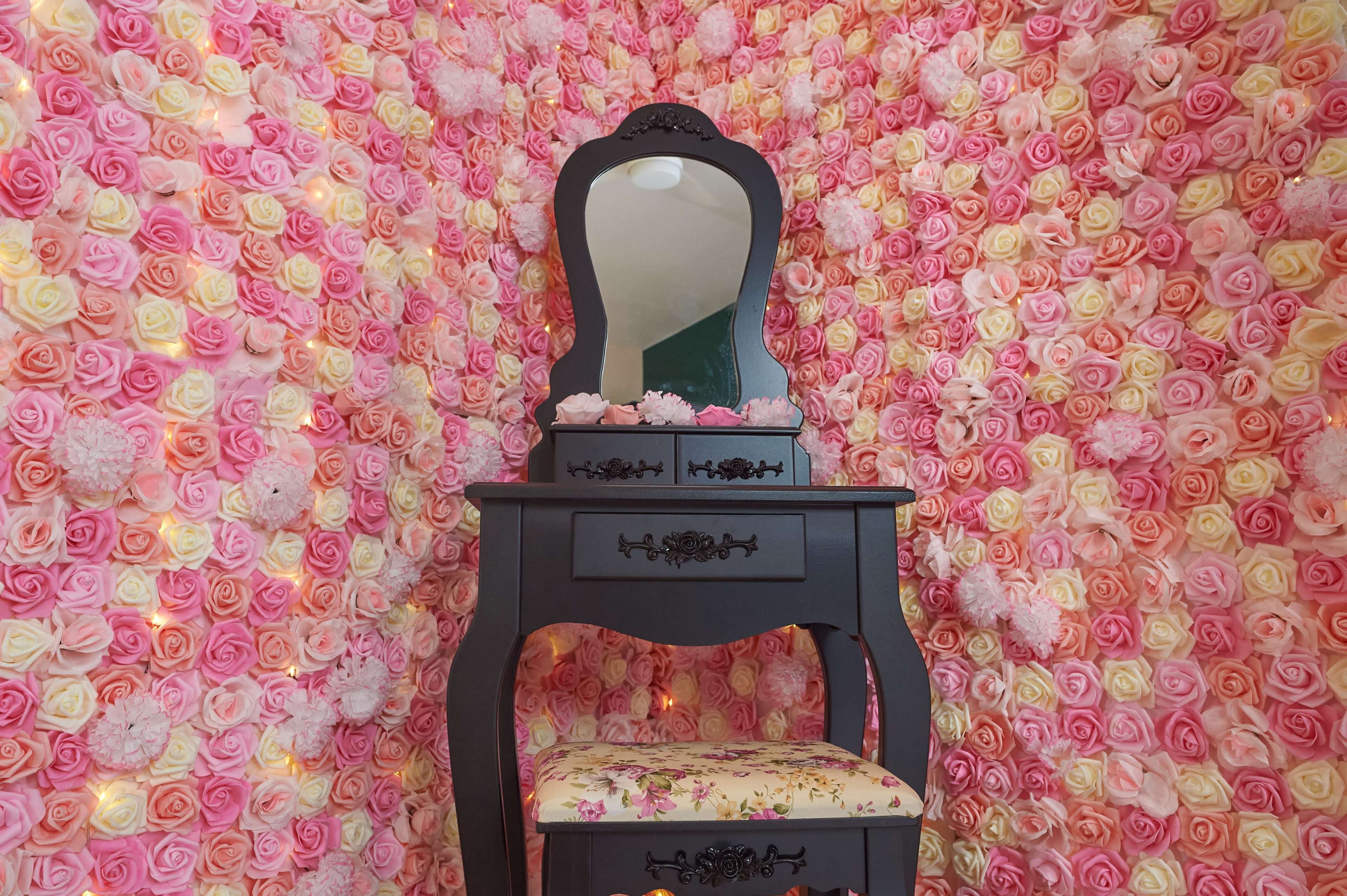 A black vanity with a mirror and a floral-patterned stool is set against a background of pink and yellow rose decorations.