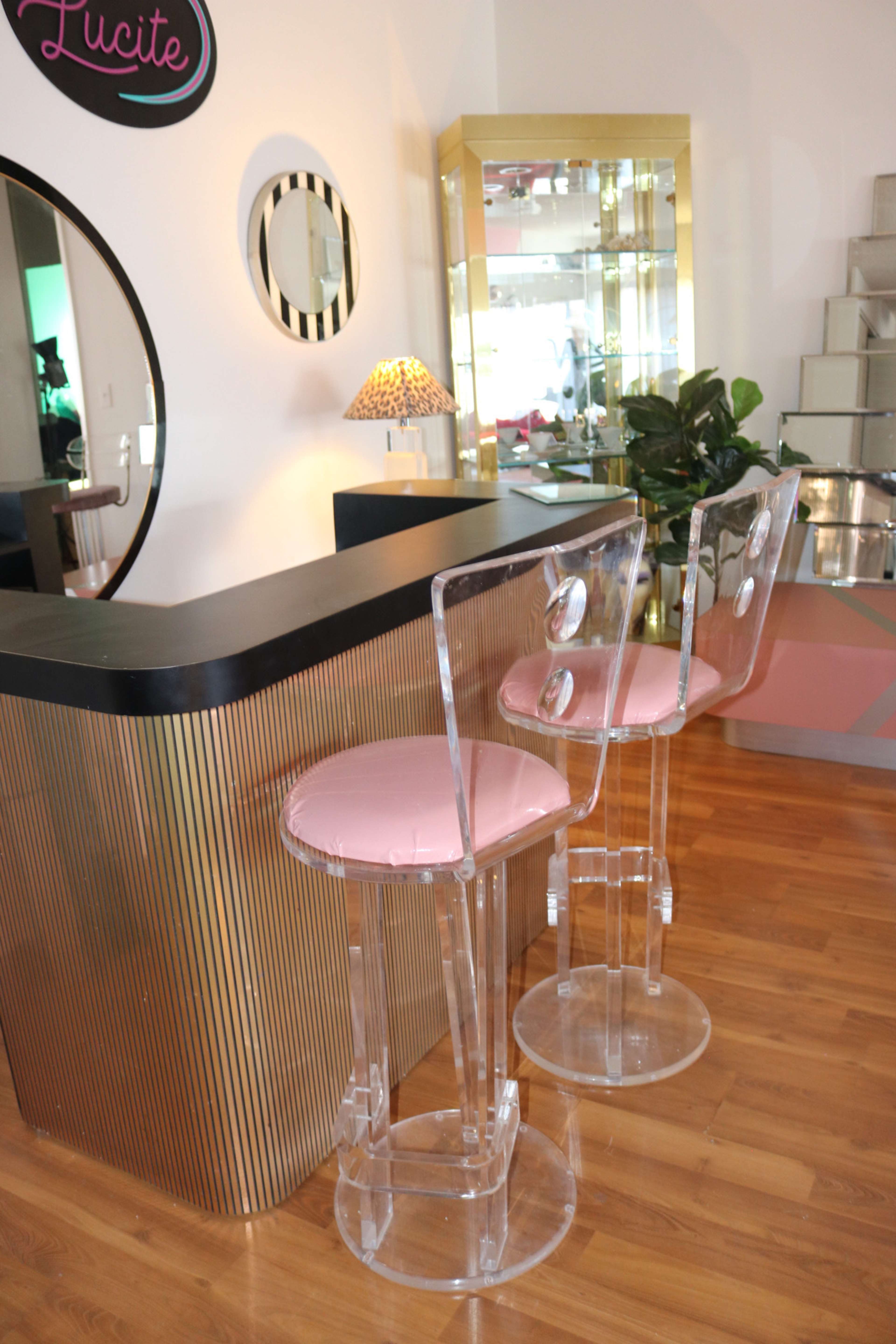 A modern bar area features two clear acrylic stools with pink seats, set against a striped counter and a large circular mirror.