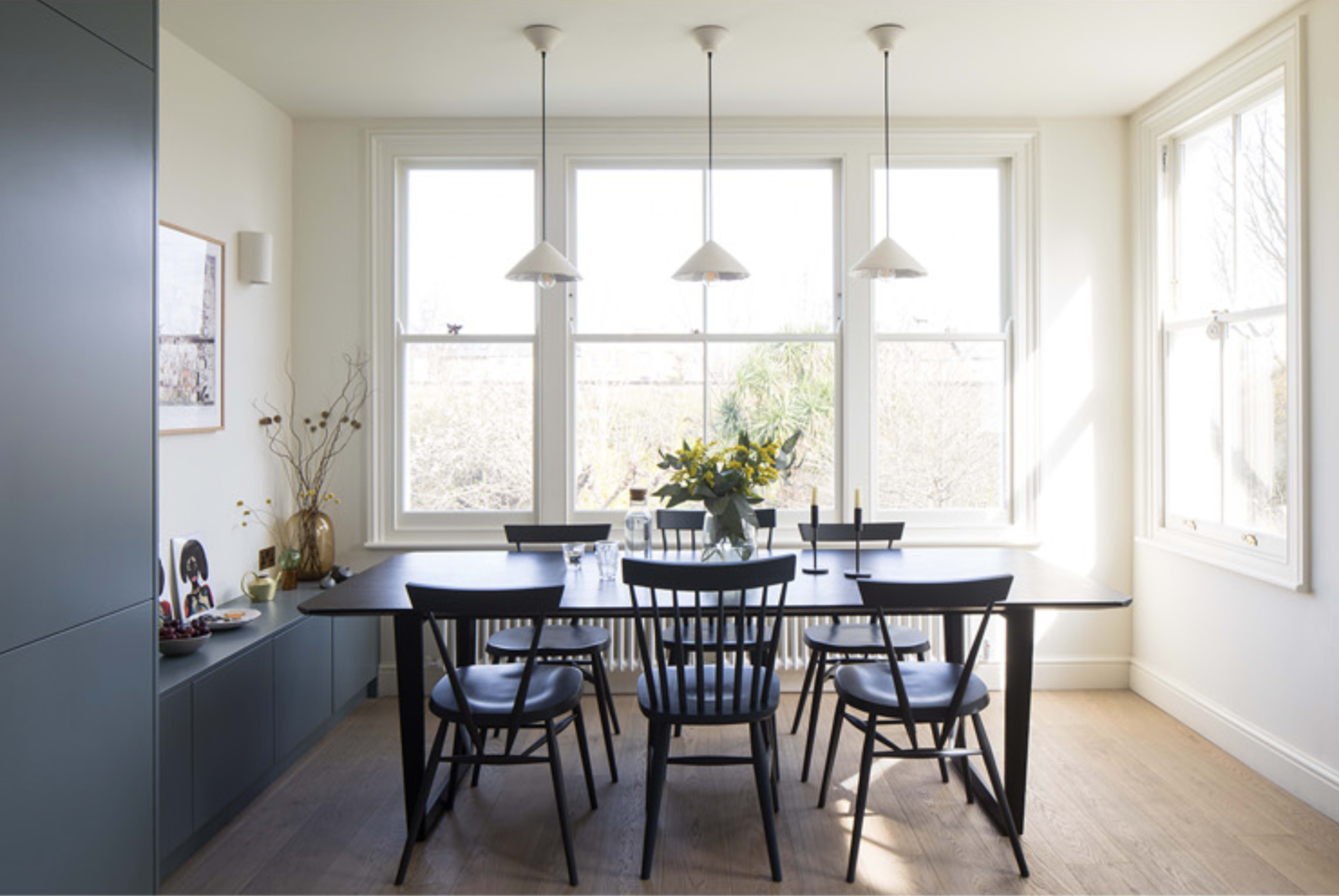 A modern dining room features a long black table surrounded by black chairs, with three pendant lights above and large windows letting in natural light.
