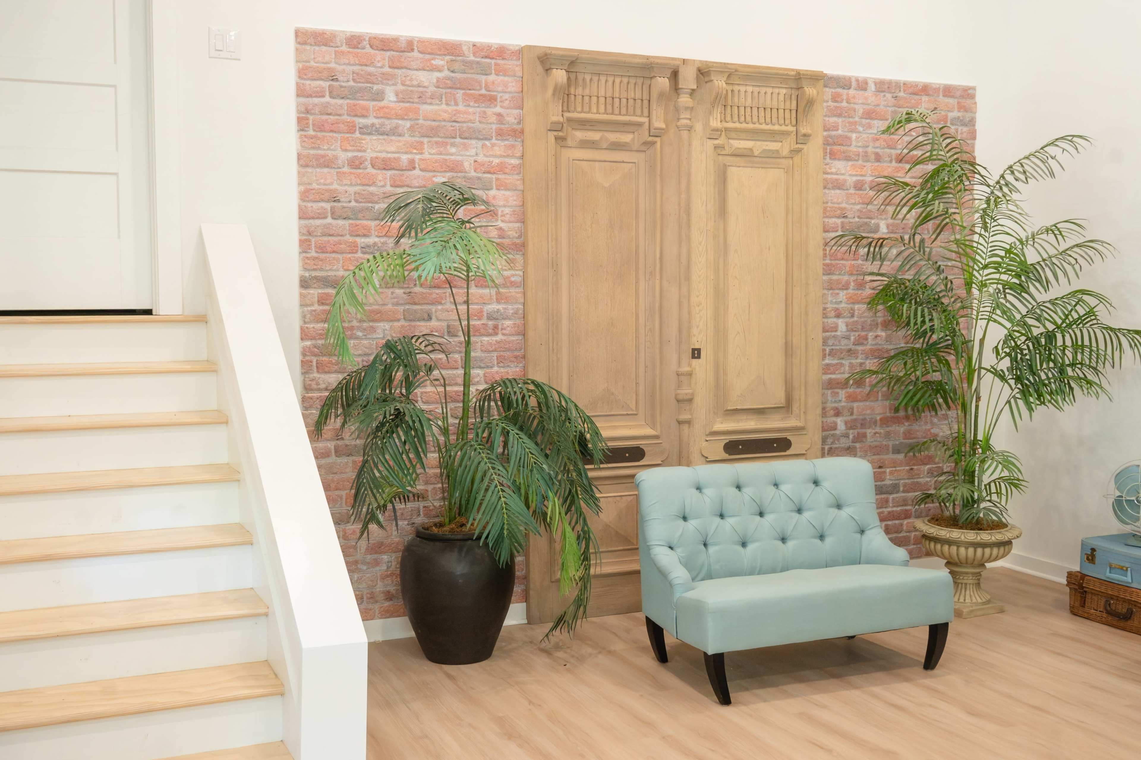 A light-colored sofa is positioned near a decorative wooden door and two potted plants against a brick-patterned wall, with a staircase visible on the left.