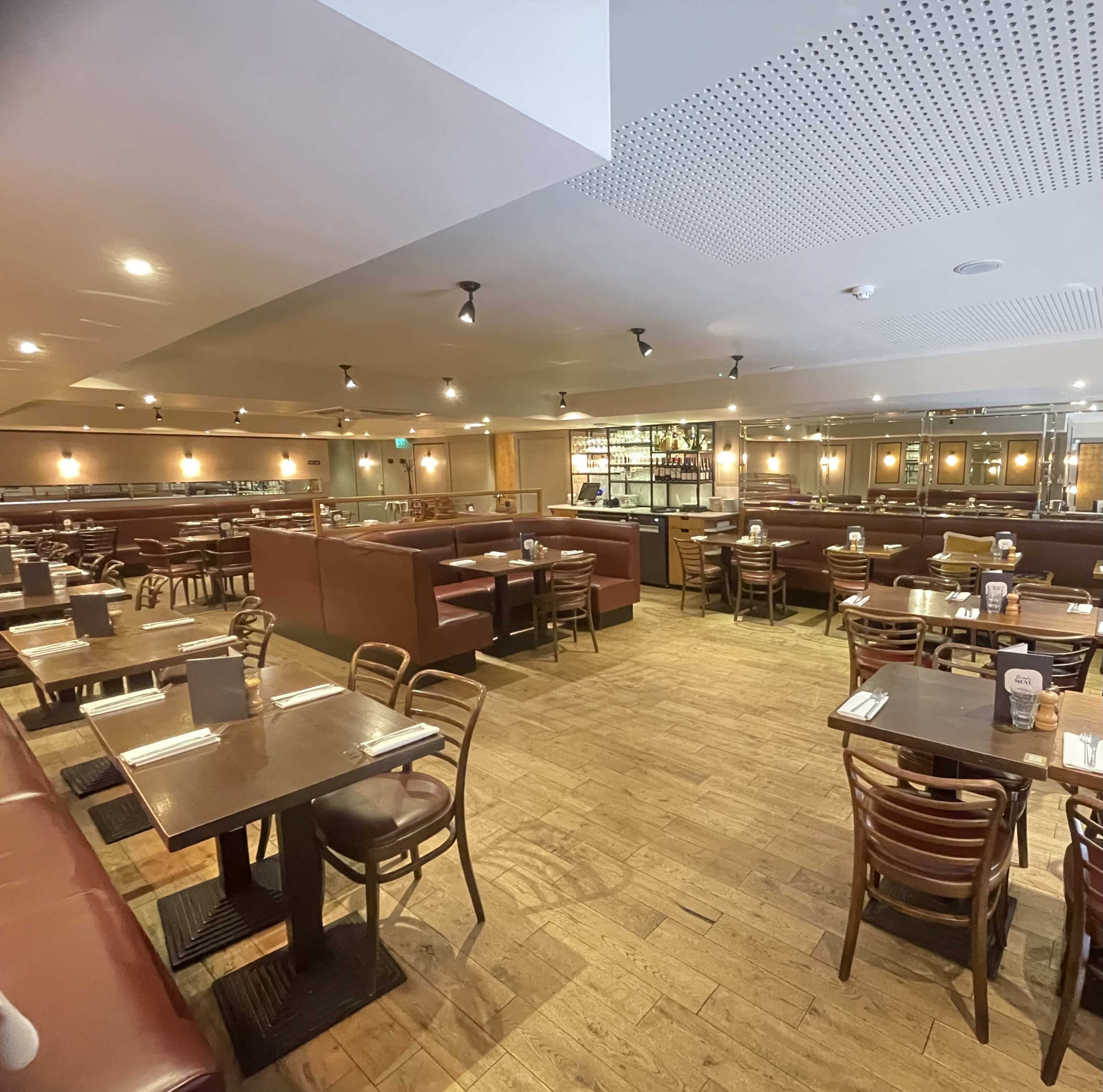 The image shows a spacious restaurant interior with wooden flooring, brown leather booths, and wooden tables set for dining.