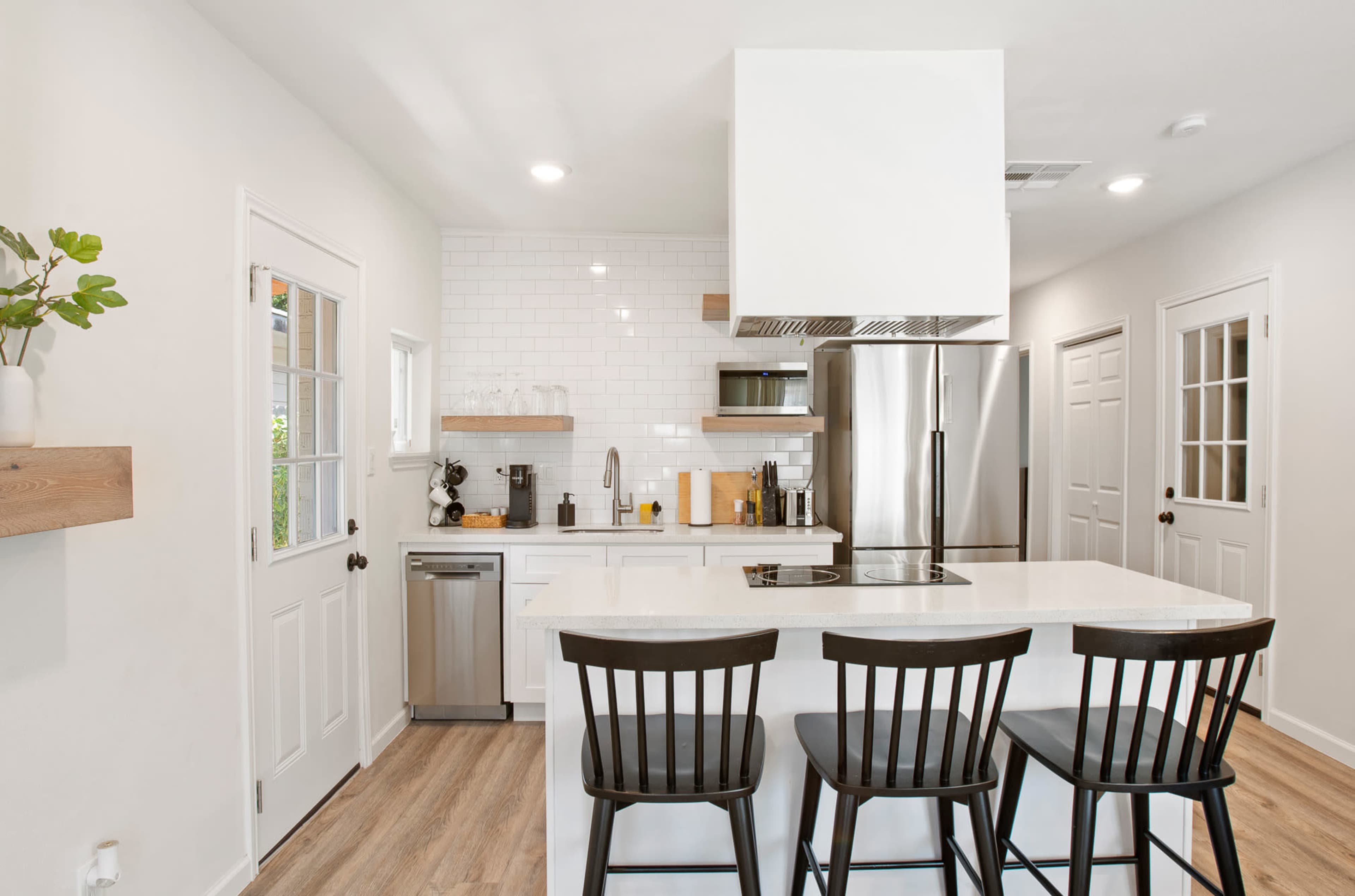 The image shows a modern kitchen with white cabinetry, a stainless steel refrigerator, and three black bar stools at a white countertop.