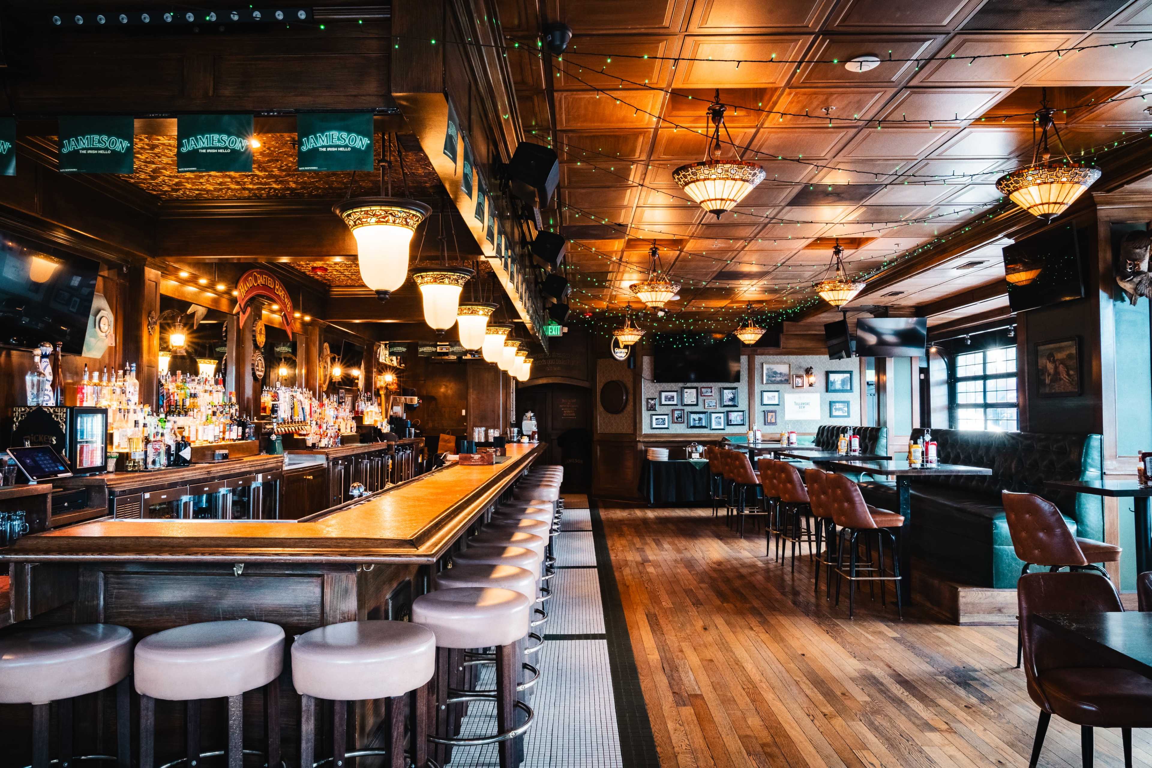 The image shows a dimly lit bar with a long wooden counter, surrounded by chairs, and decorated with hanging lights and wall photographs.