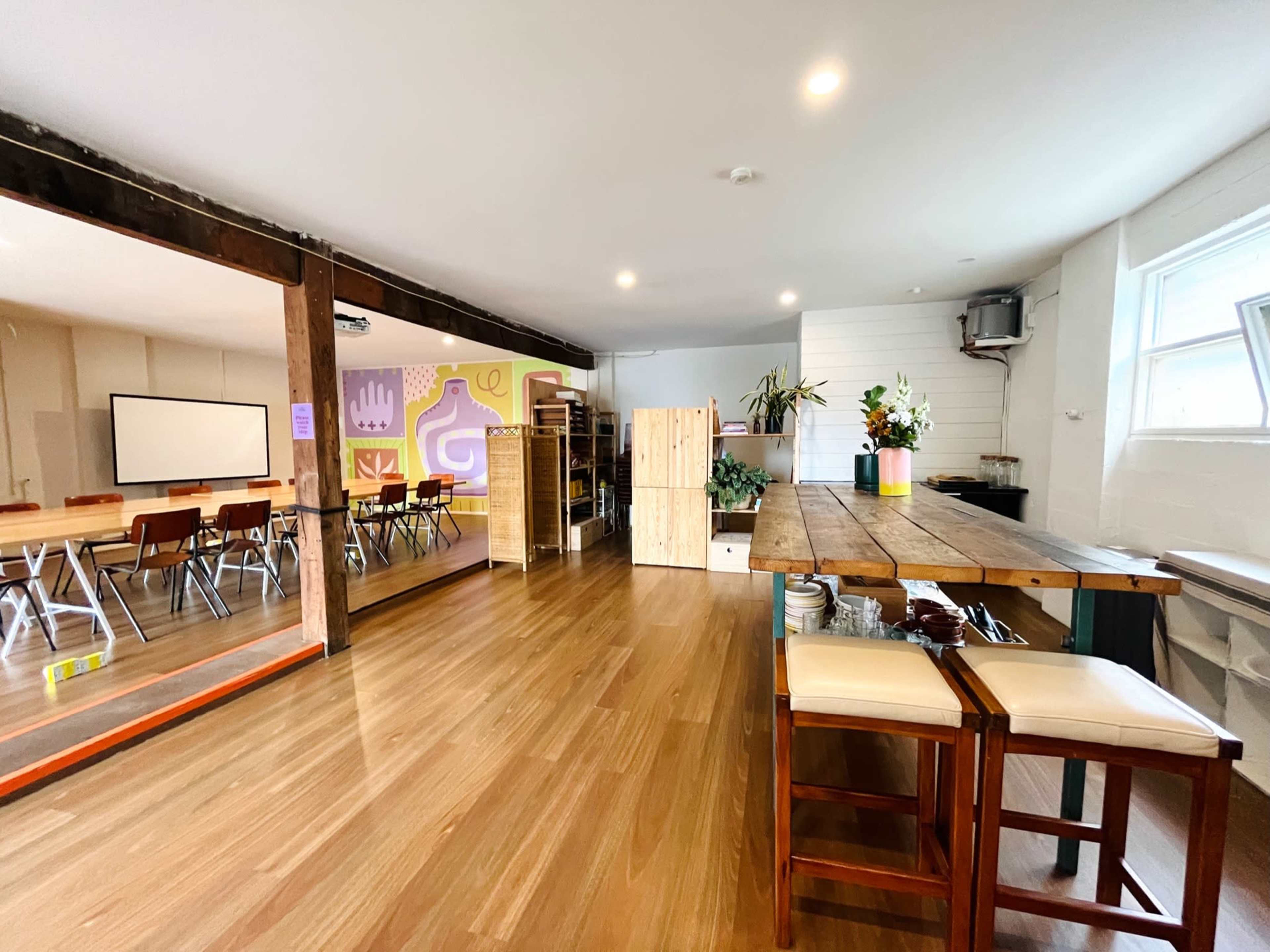 The image shows a spacious, well-lit classroom and work area with a dining table, wooden furniture, and colorful wall art.