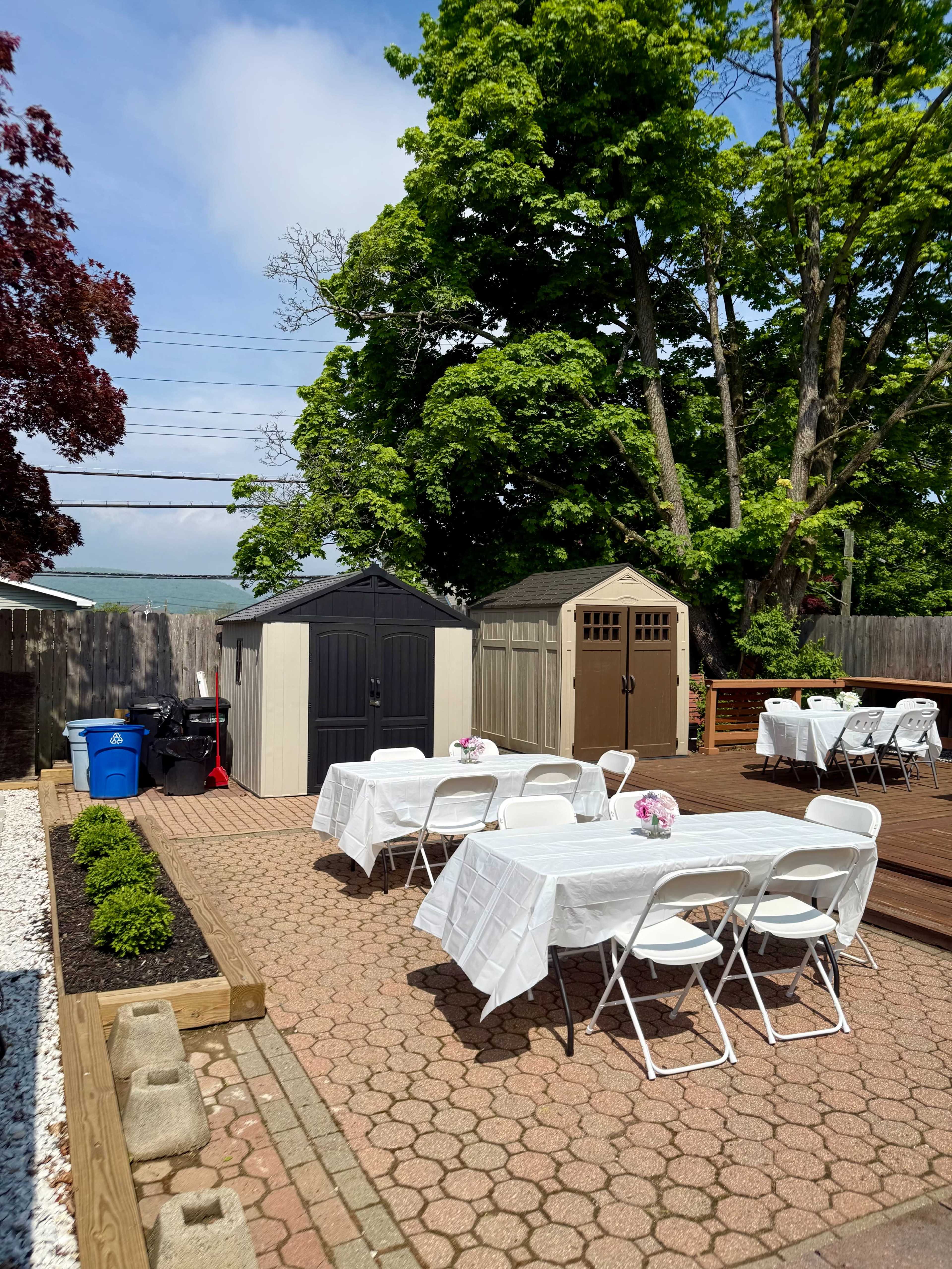 The image shows a landscaped outdoor area with two storage sheds, several white tables set with tablecloths, and a view of trees and a blue sky in the background.
