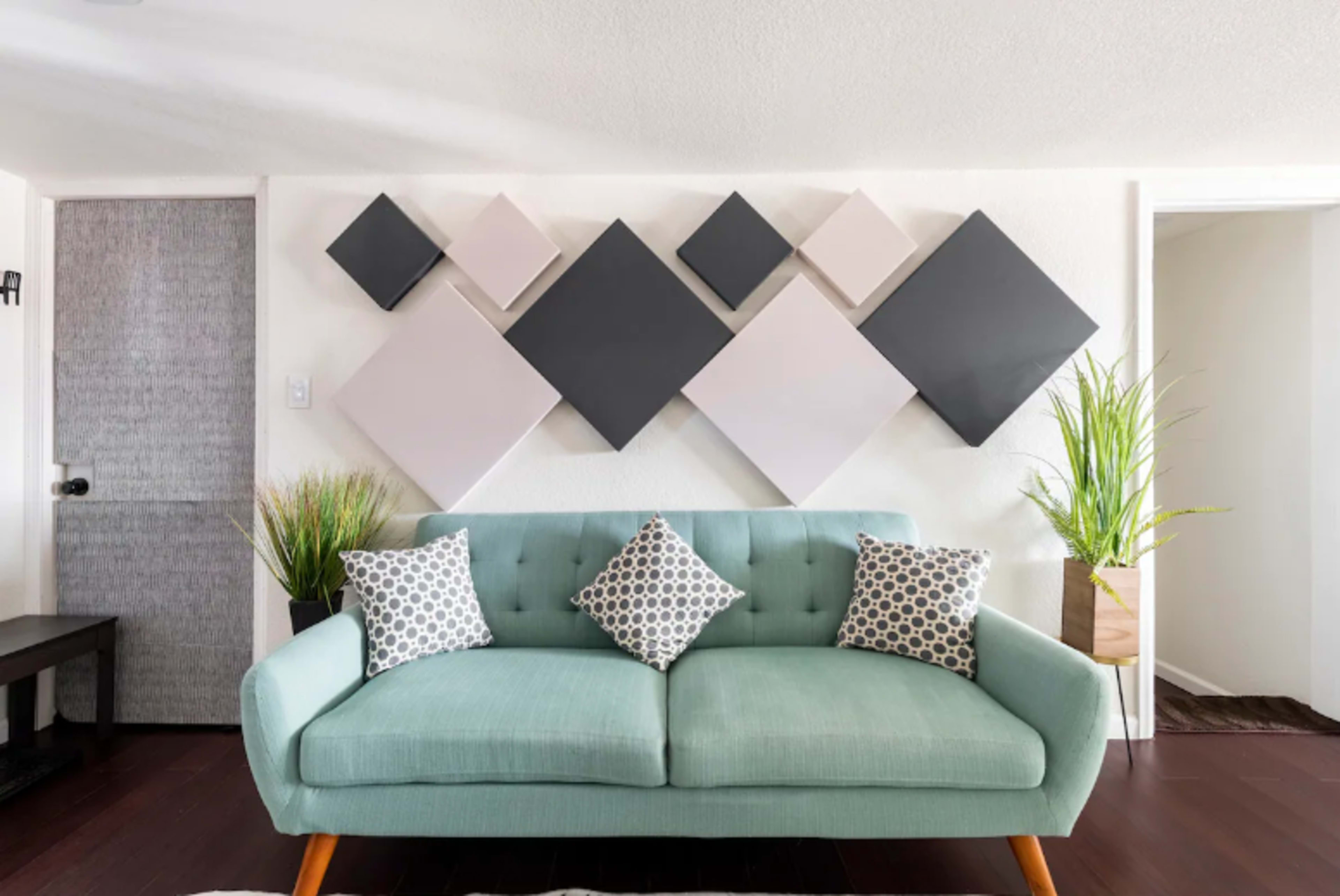 A light teal sofa with patterned pillows is positioned in front of a wall featuring an arrangement of geometric wall art and potted plants.
