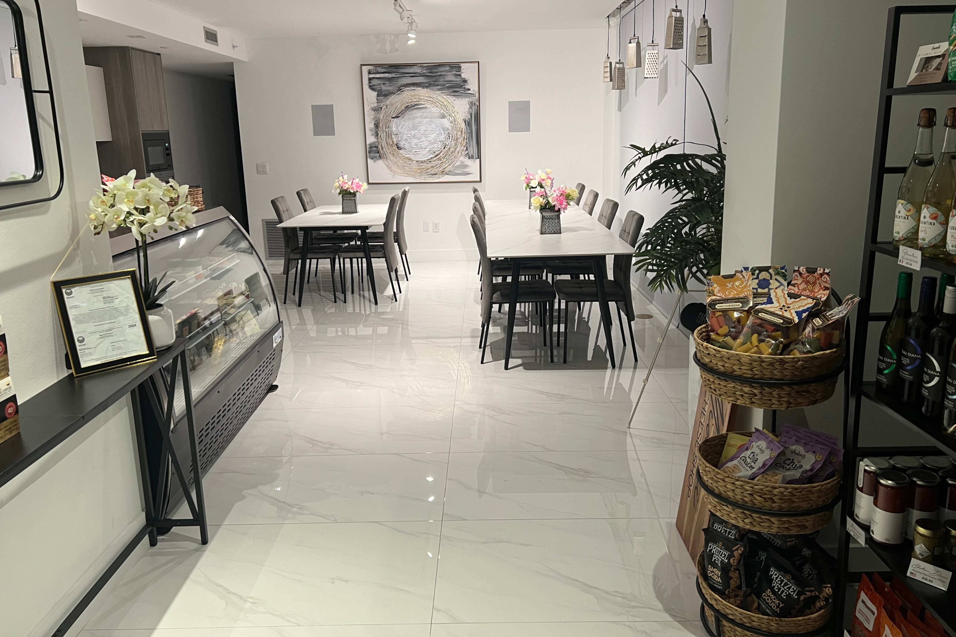 A modern interior of a café with several tables and chairs arranged around a display case and a shelving unit filled with snacks and beverages.