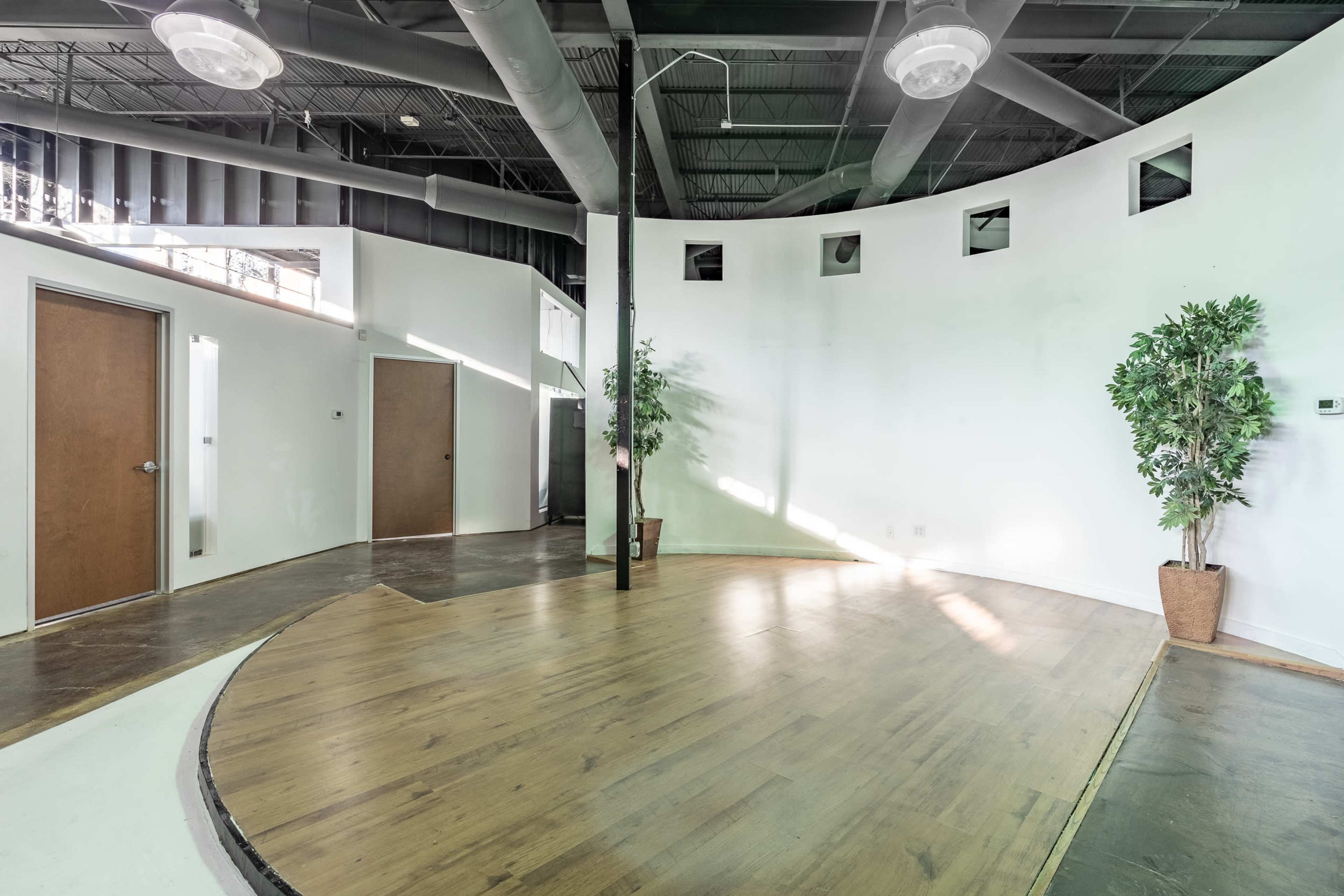 The image shows a spacious, partially furnished room with curved walls, wooden flooring, and two doors leading to adjacent spaces.