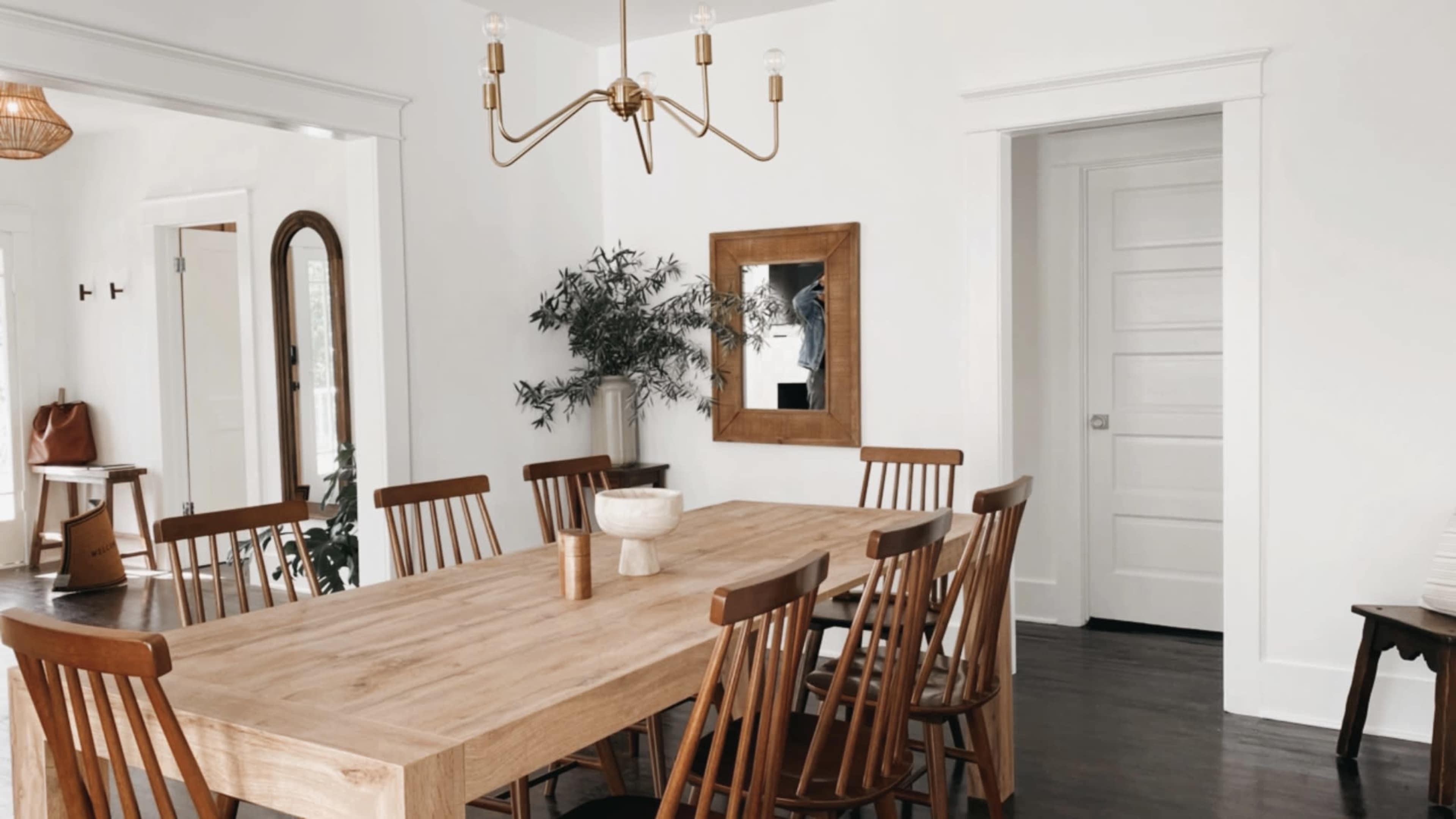 A wooden dining table with ten chairs is positioned in a bright room featuring a plant, a mirror, and a light fixture.