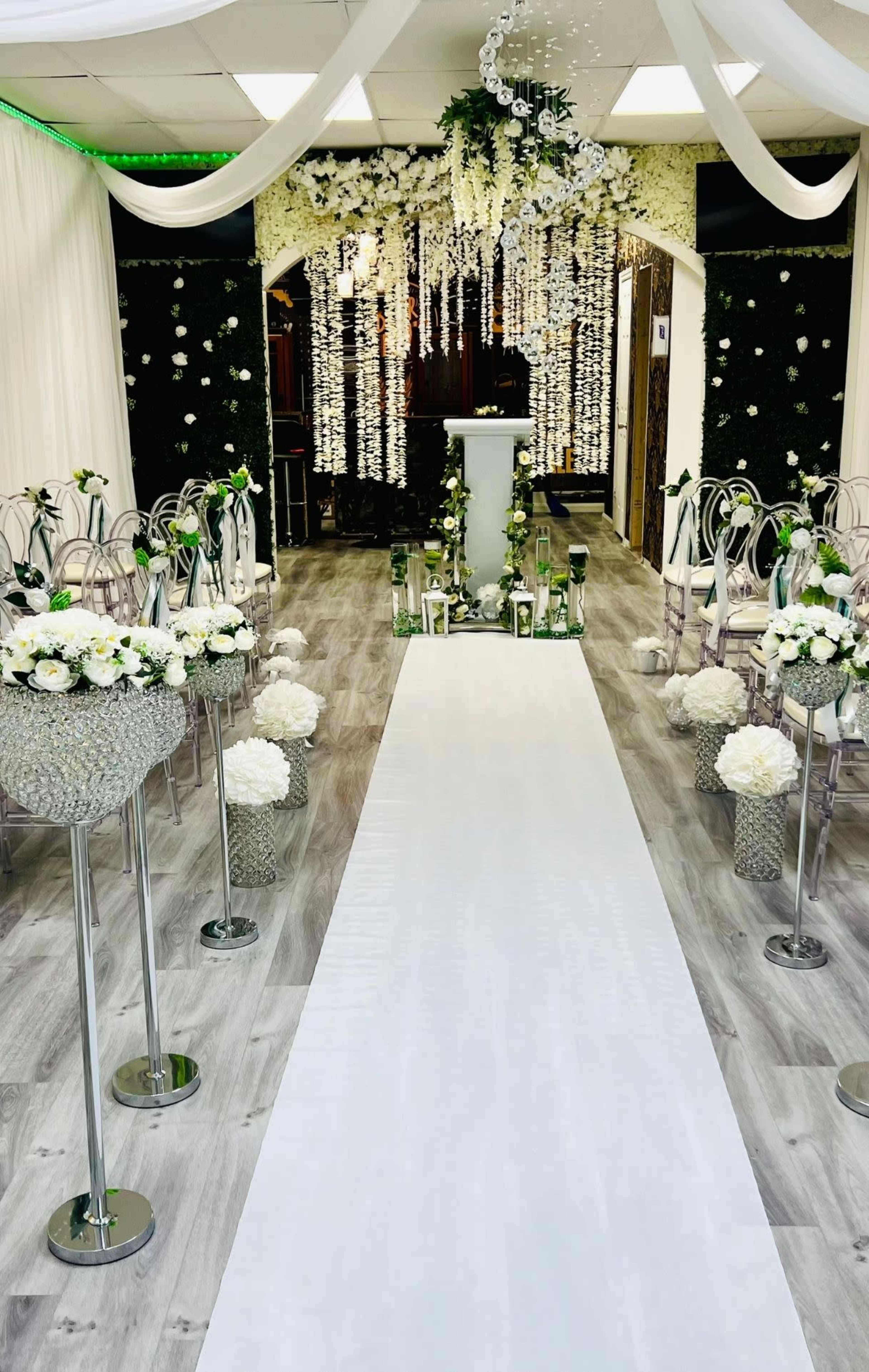 A decorated indoor space features a white aisle leading to an altar surrounded by flower arrangements and hanging decorations.