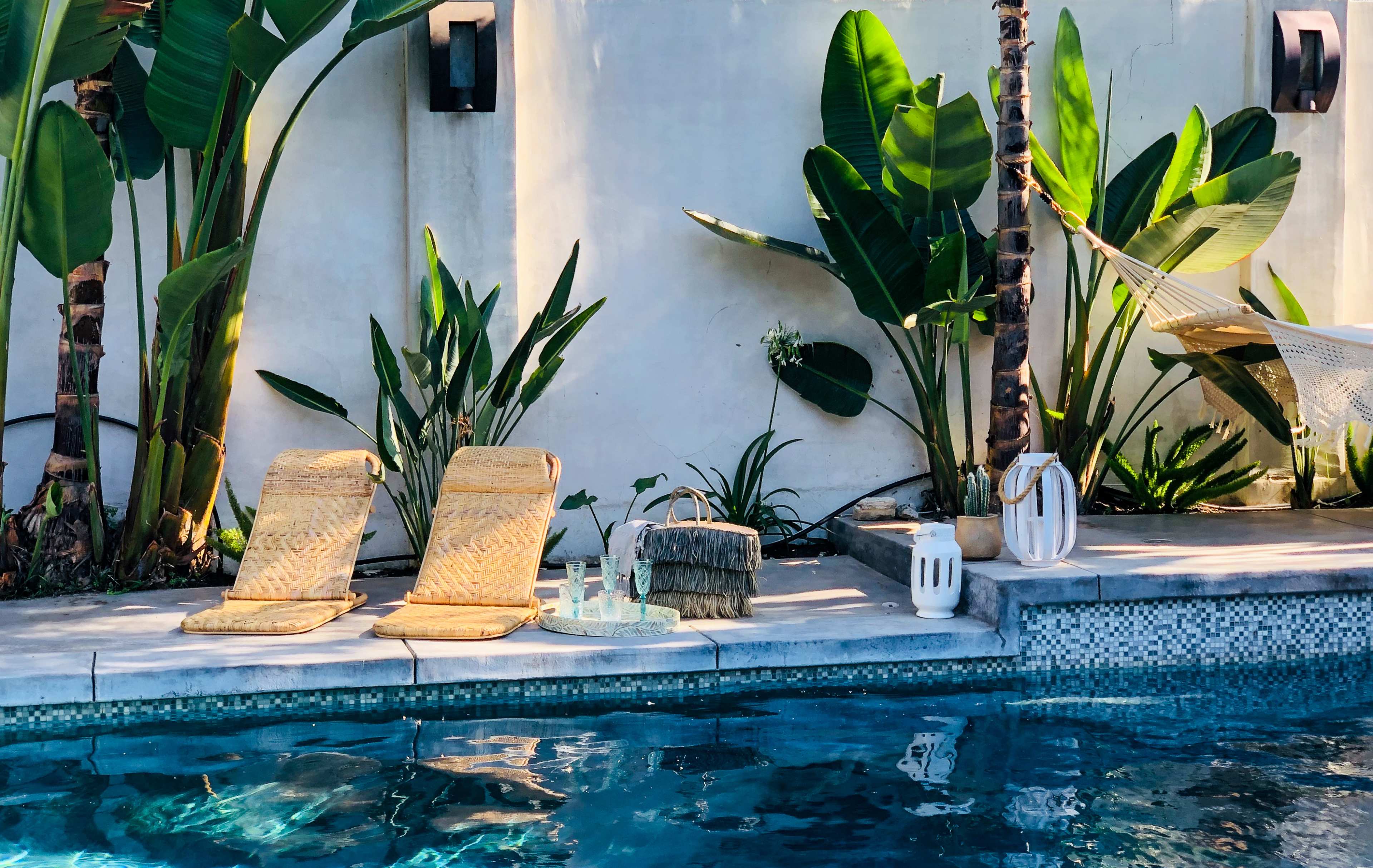 A calm poolside scene features two yellow lounge chairs, a decorative basket, and various plants near the water.