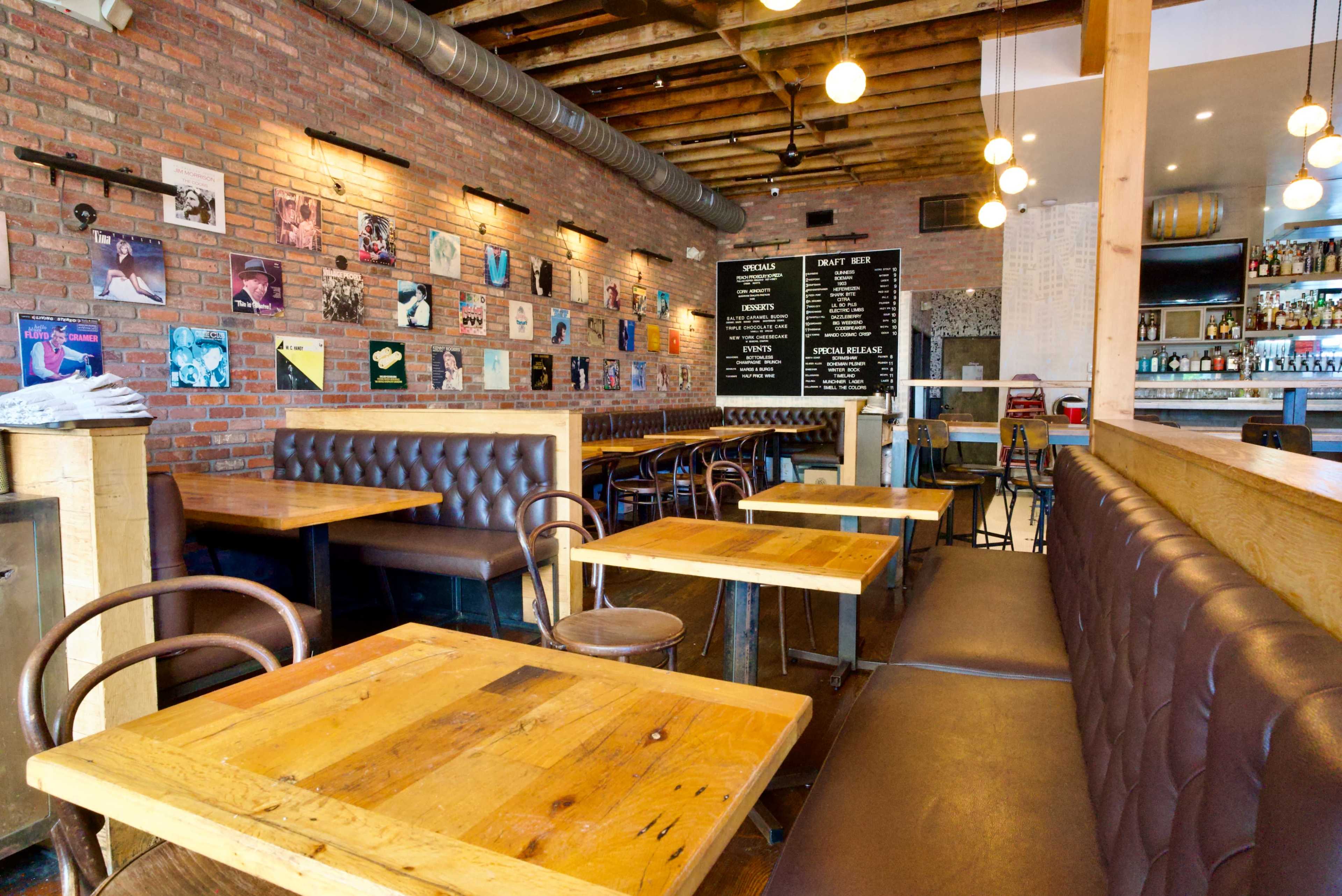The image shows a trendy restaurant interior with wooden tables, leather benches, a brick wall featuring vinyl records, and a bar area in the background.