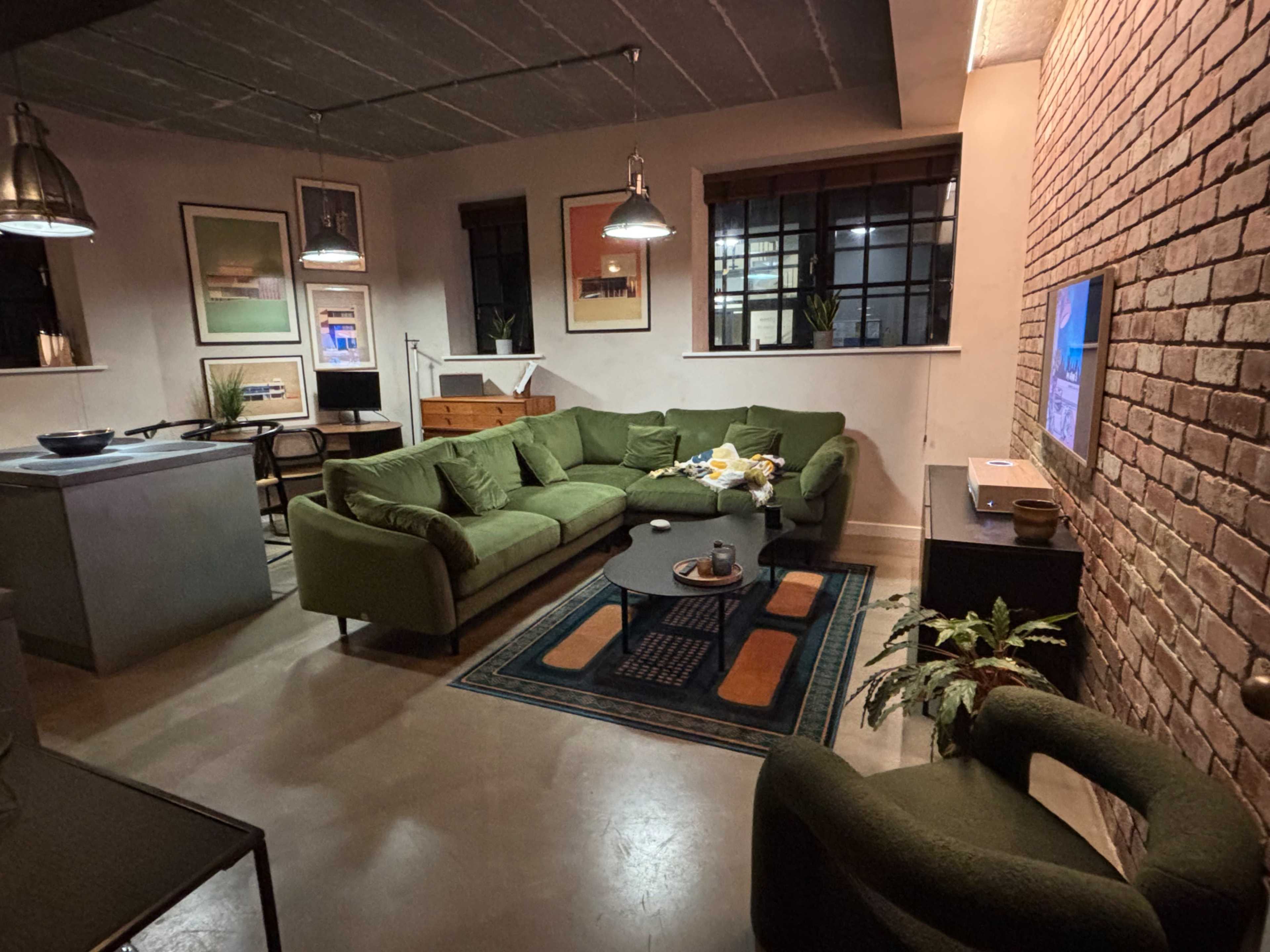 The image shows a modern living room with a green sectional sofa, a coffee table, and brick walls, illuminated by hanging lights and natural window light.