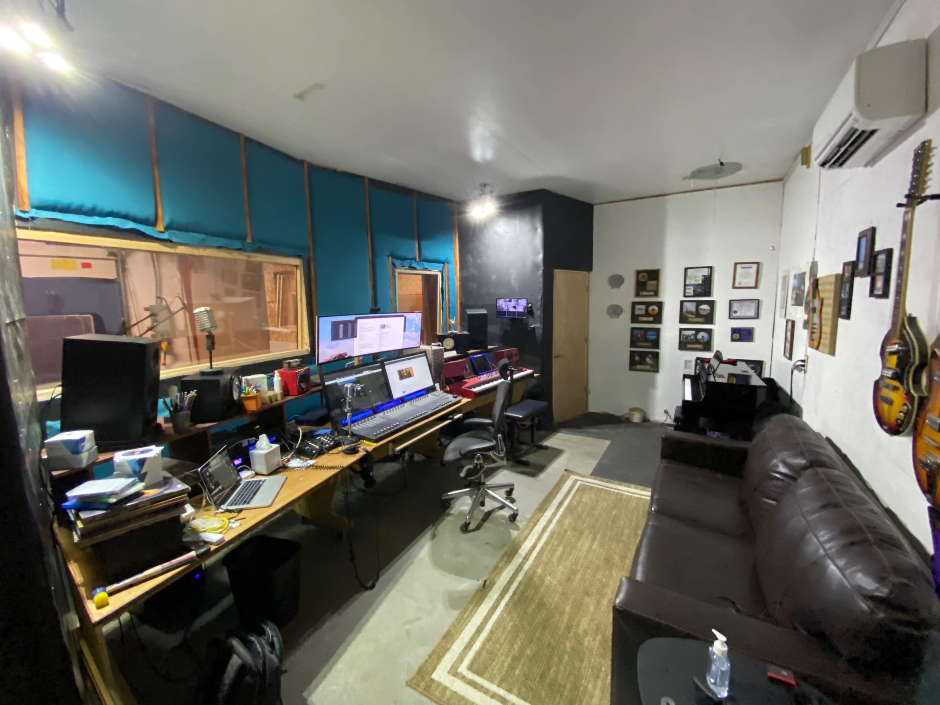 The image shows a recording studio with multiple computer workstations, a leather couch, and framed photographs on the wall.