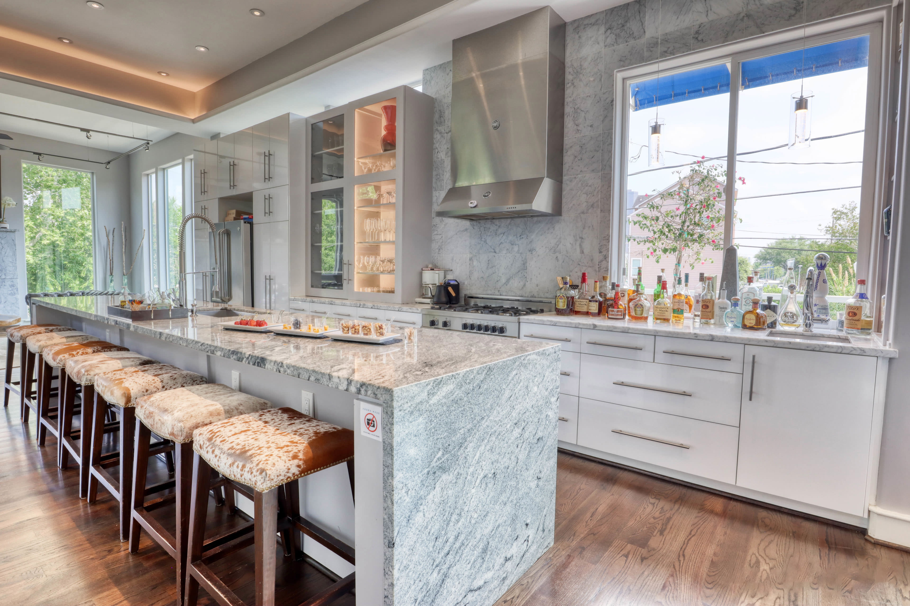 A modern kitchen features a long marble island with bar stools, stainless steel appliances, and large windows with a view of the outdoors.