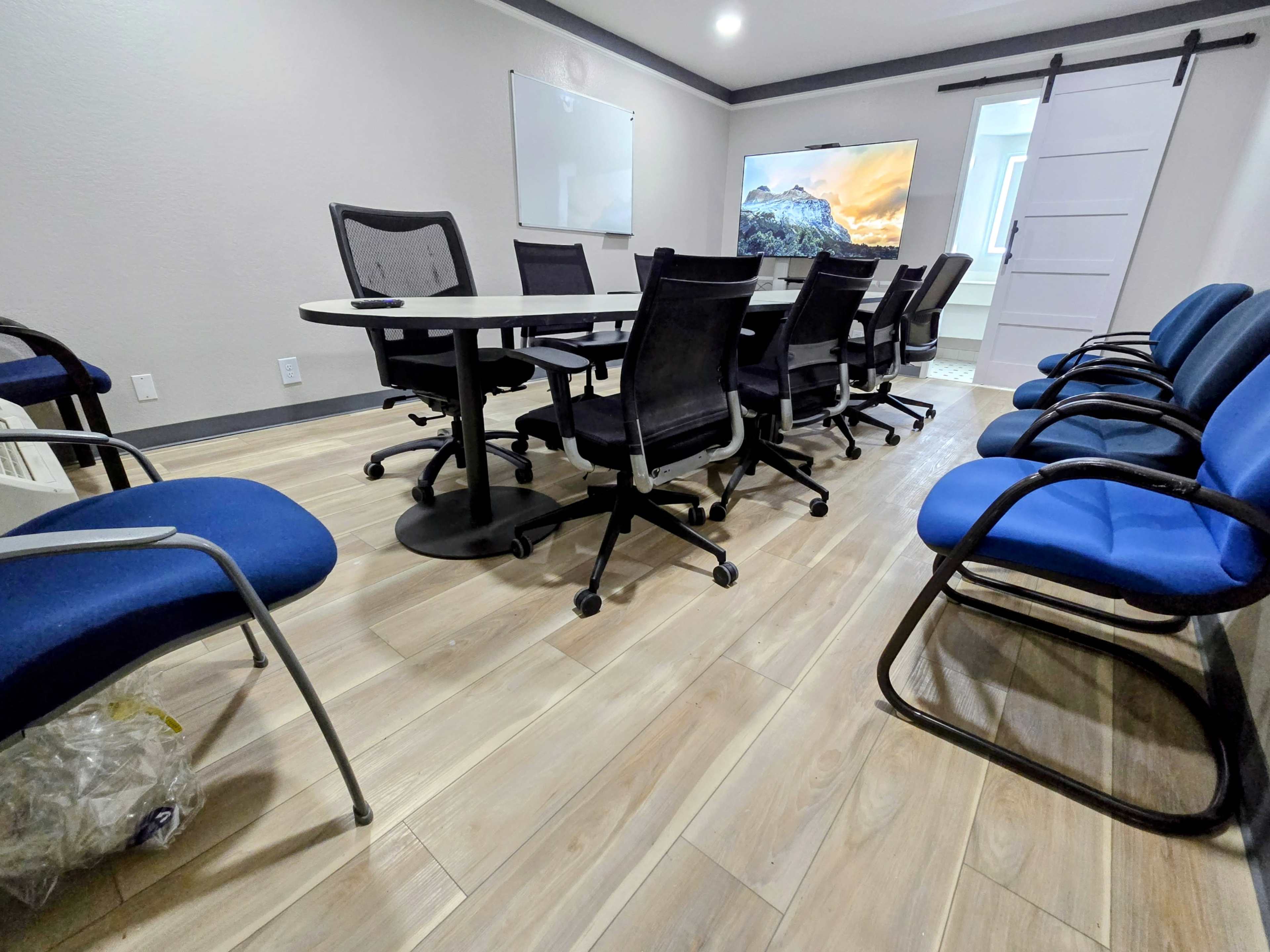 The image shows a modern conference room with a long table surrounded by black and blue chairs, a large screen mounted on the wall, and a whiteboard.