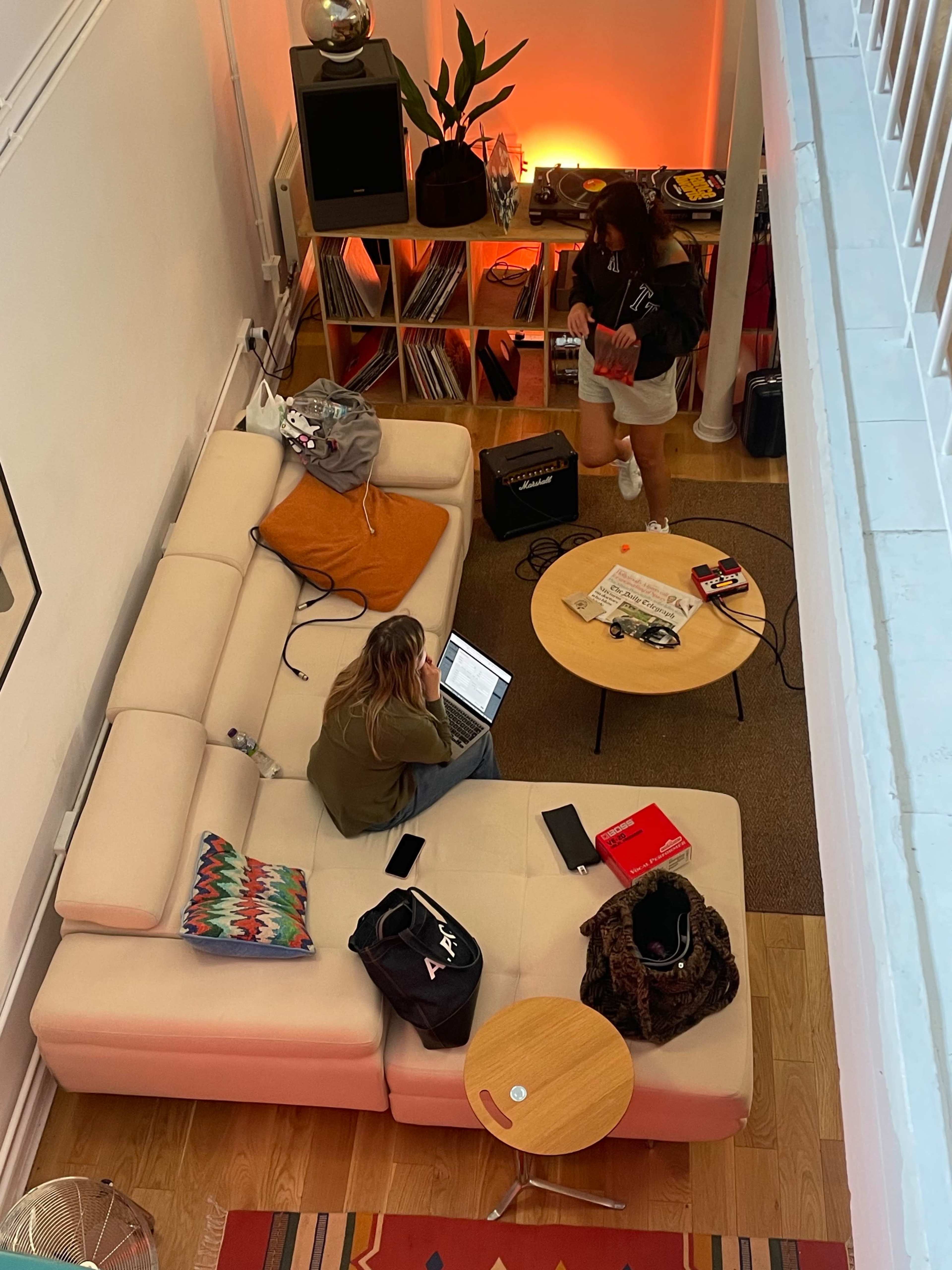 Two people in a cozy room with a white sofa, a small round table, and various items, including a guitar amplifier and records, arranged around them.