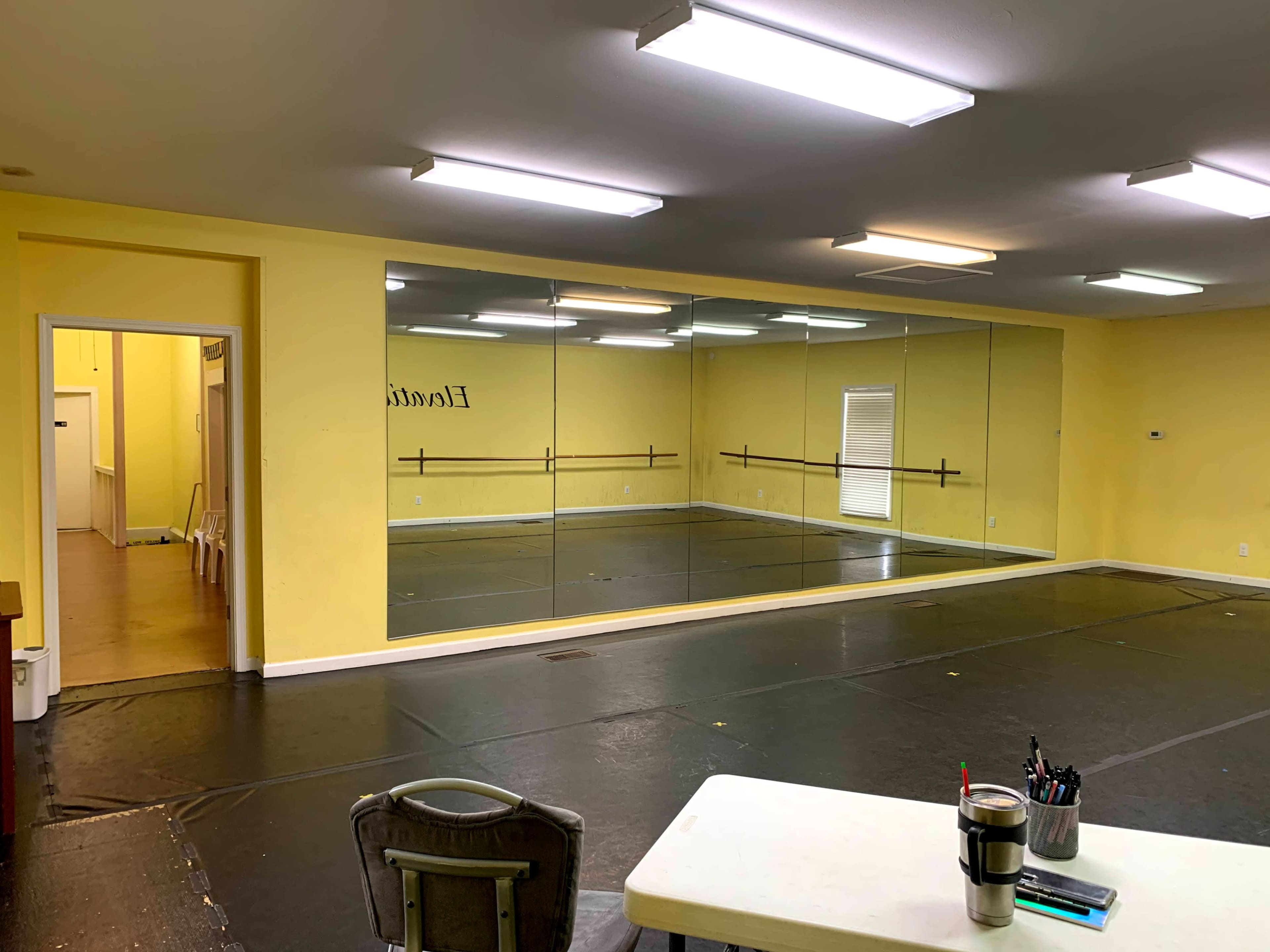 A spacious studio with yellow walls features a large mirror and a black floor, complemented by a table and chairs.