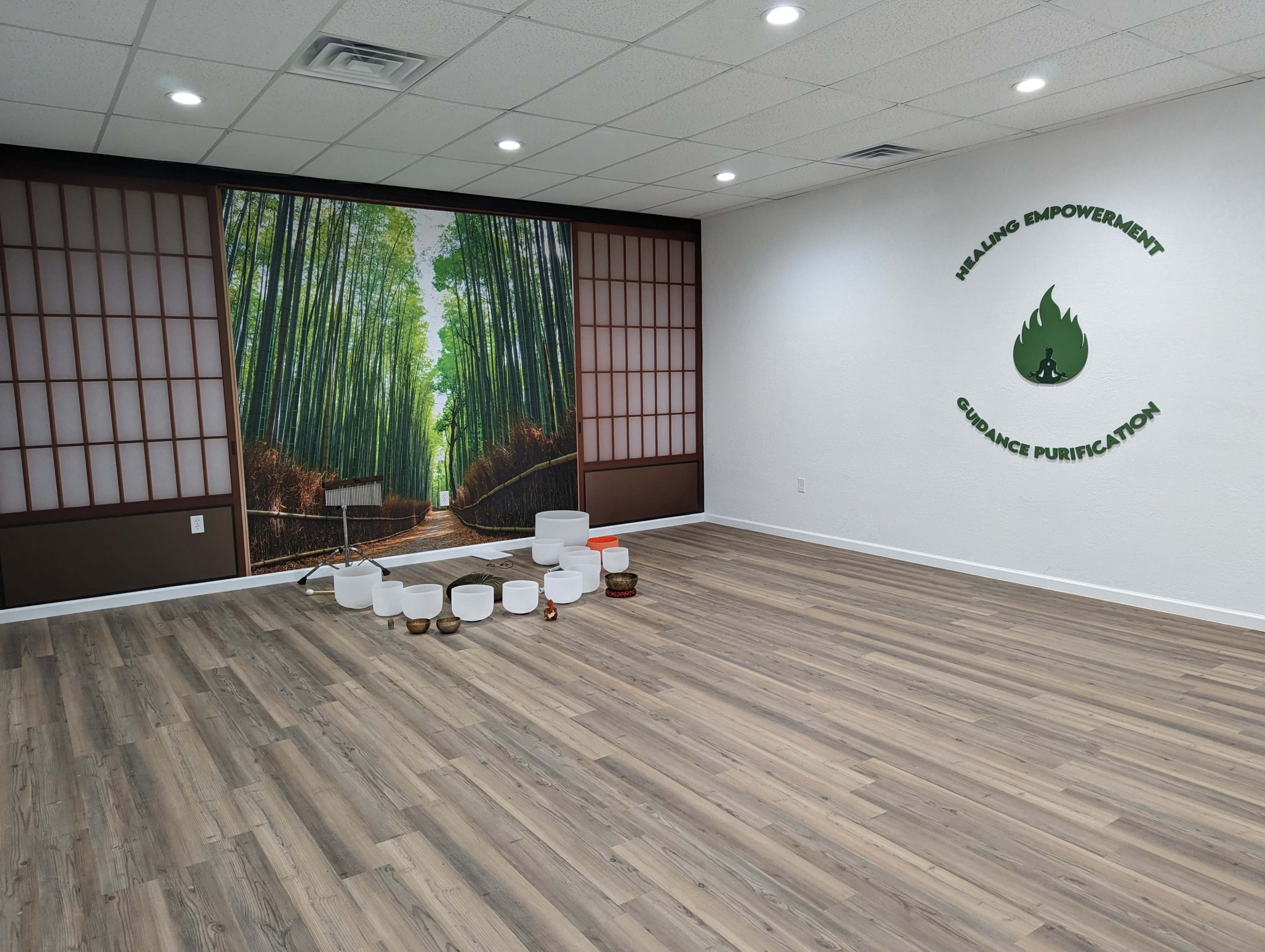A spacious room with bamboo wall art, wooden flooring, and various sound healing bowls arranged on the floor.