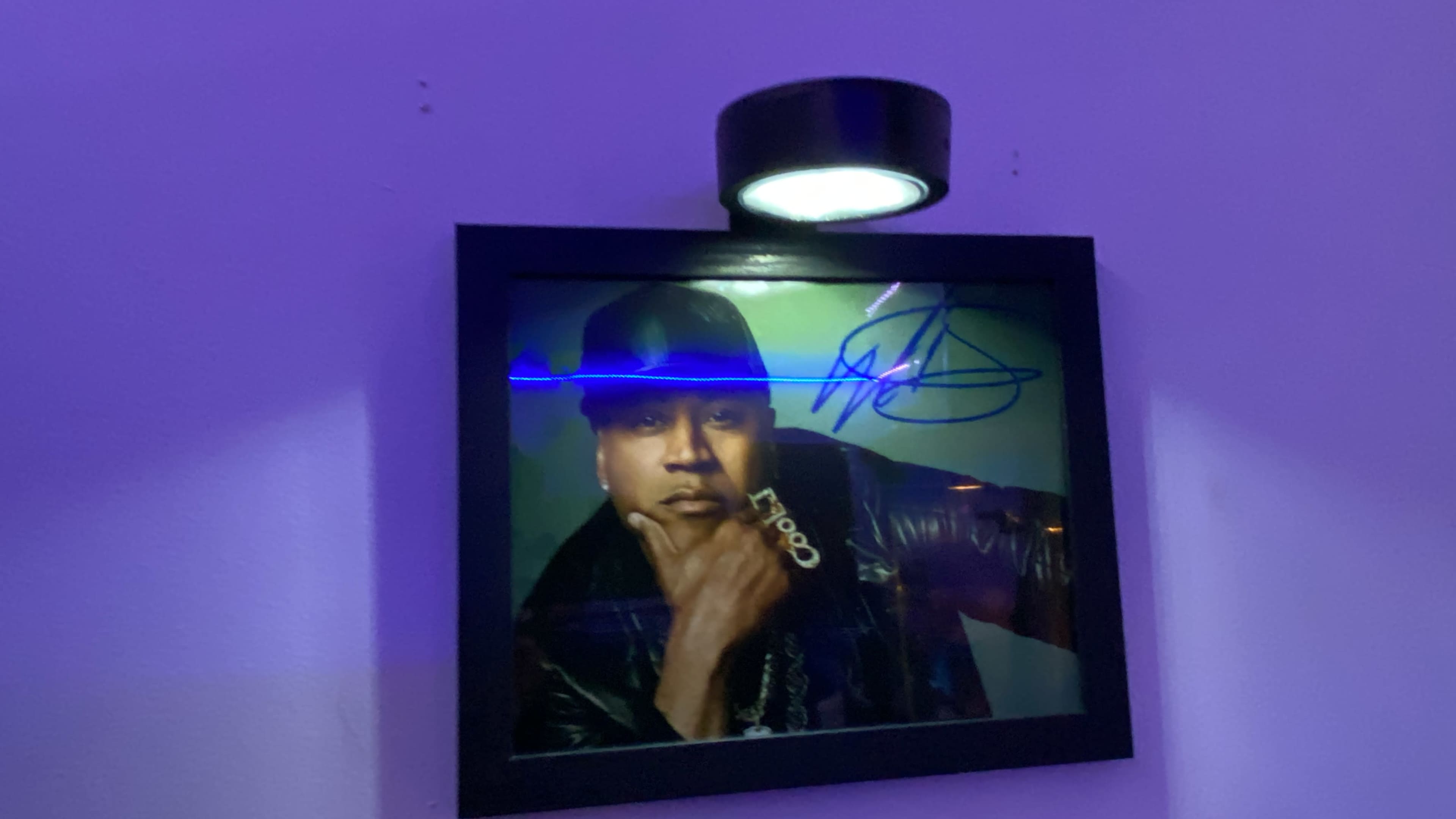 A framed, autographed portrait of a man in a black hat and jacket, illuminated by a mounted light on a purple wall.
