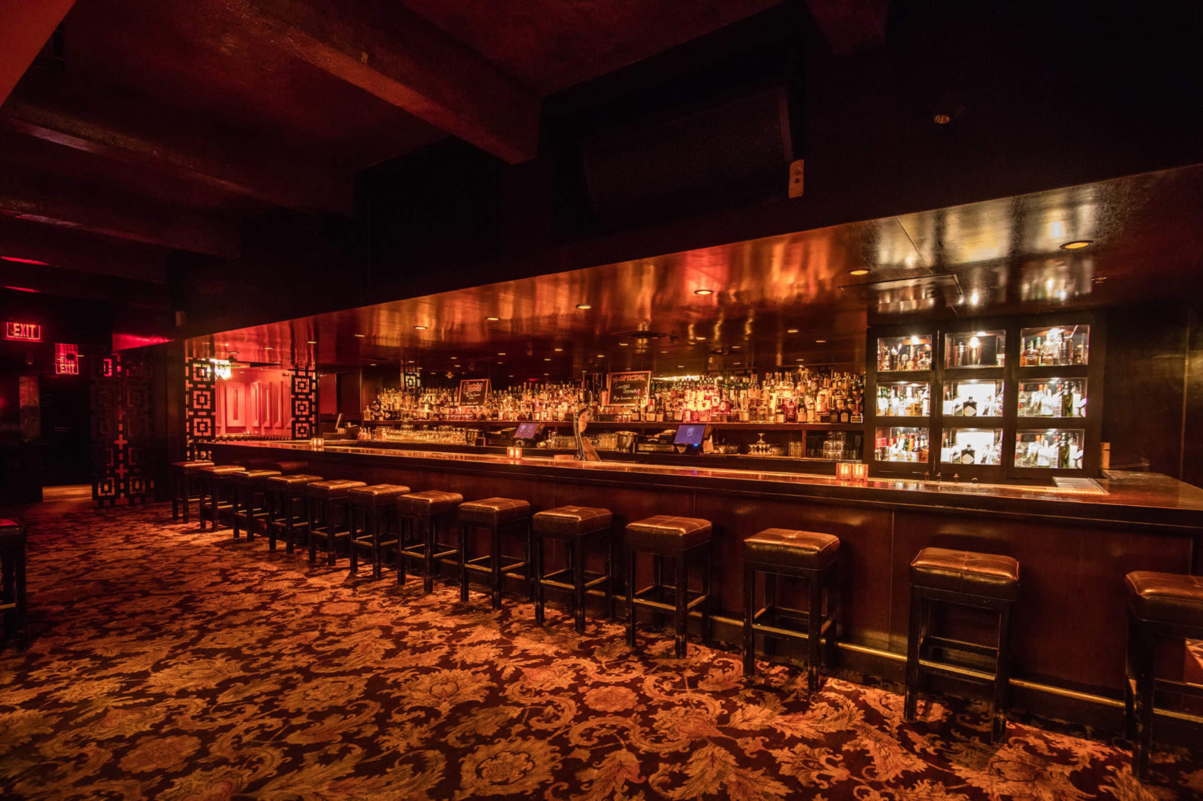 The image shows a dimly lit bar with a long wooden counter, lined with barstools, and shelves displaying various bottles of liquor against a dark backdrop.