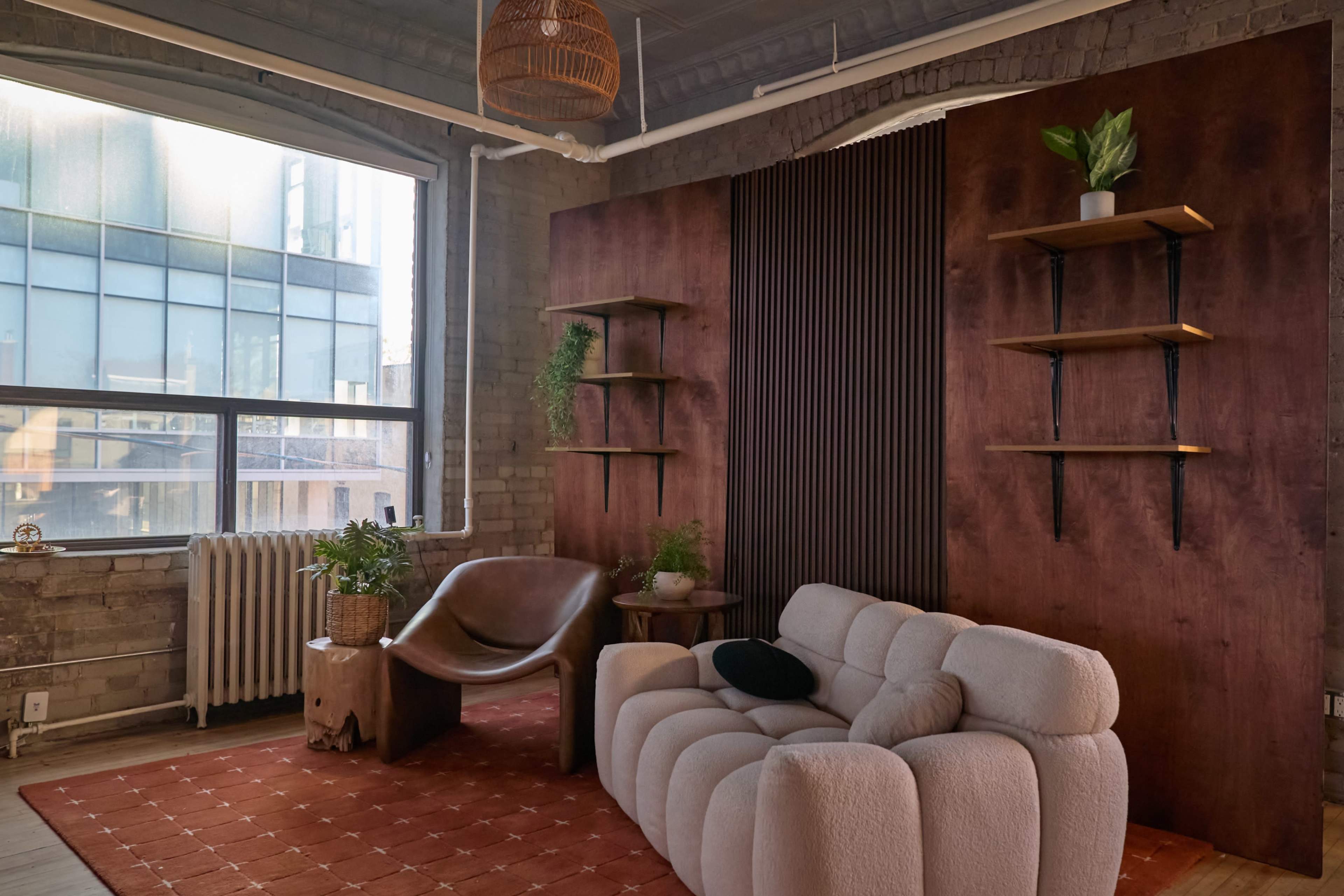 The image shows a modern living room with a curved beige sofa, a wooden accent wall with shelves, plants, and large windows that let in natural light.