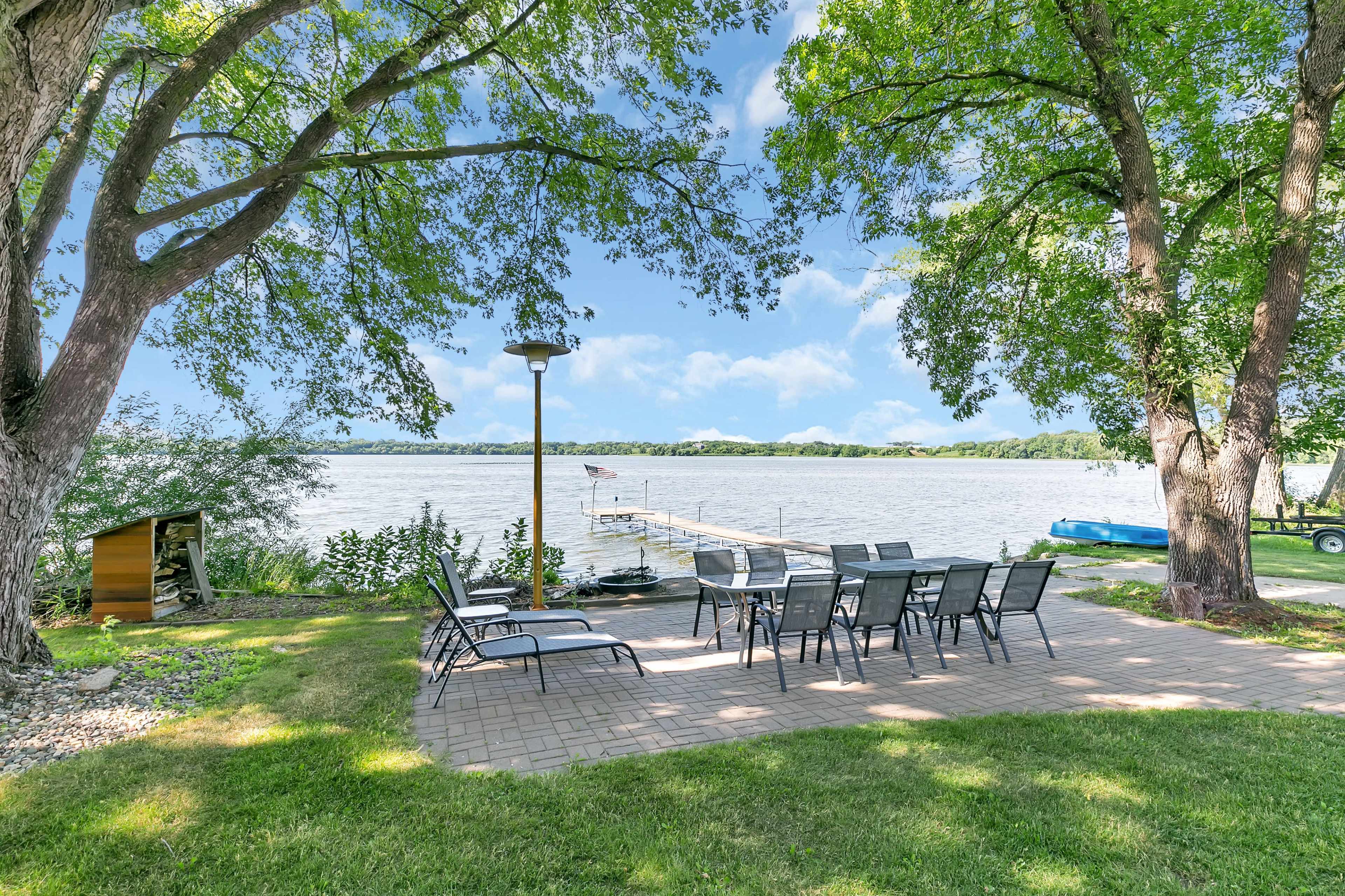 A patio area with outdoor seating and a lamp overlooks a lake, flanked by trees and a dock.