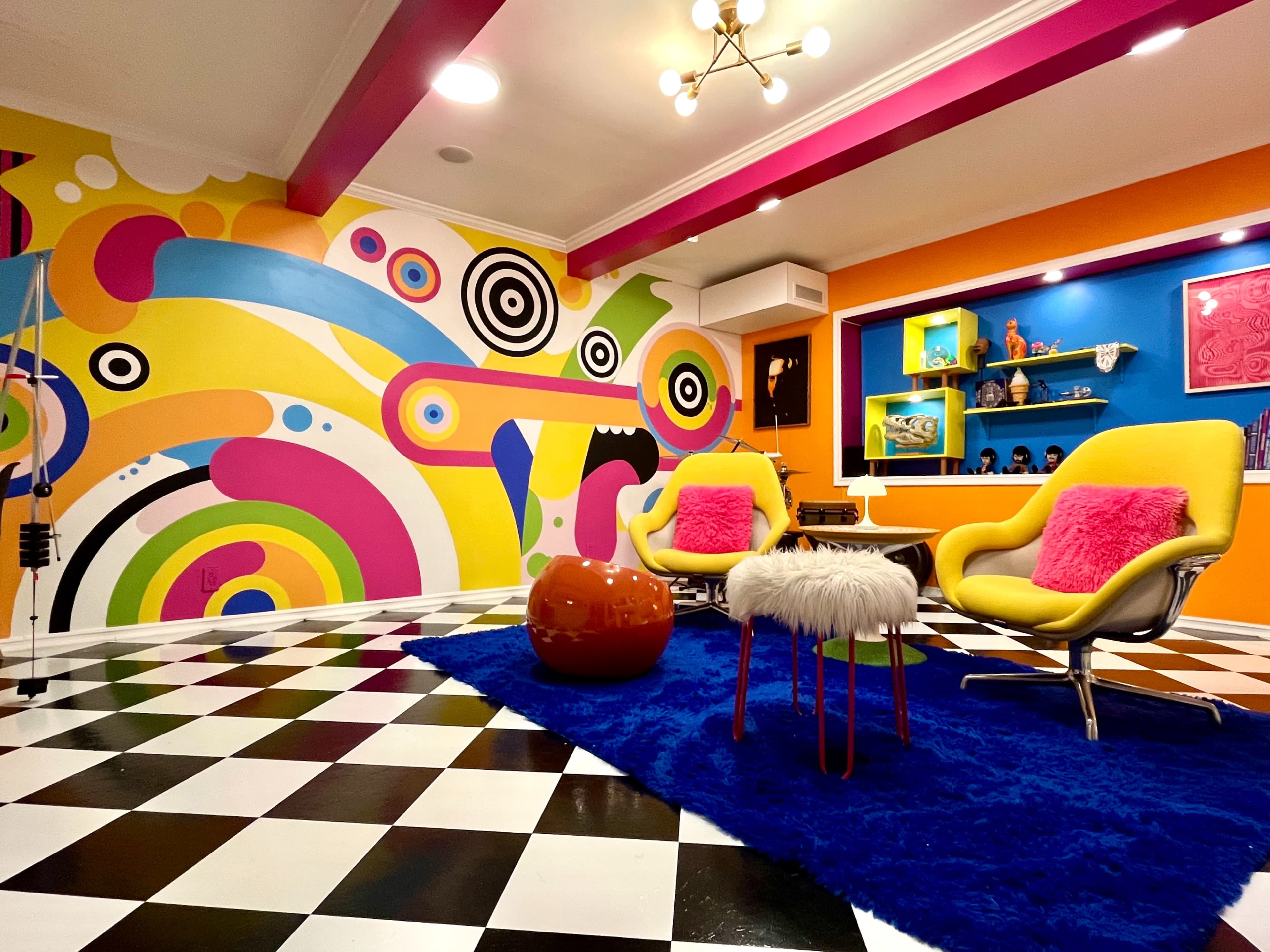 The image shows a brightly colored room featuring a patterned wall mural, vibrant furniture, and a checkered floor.