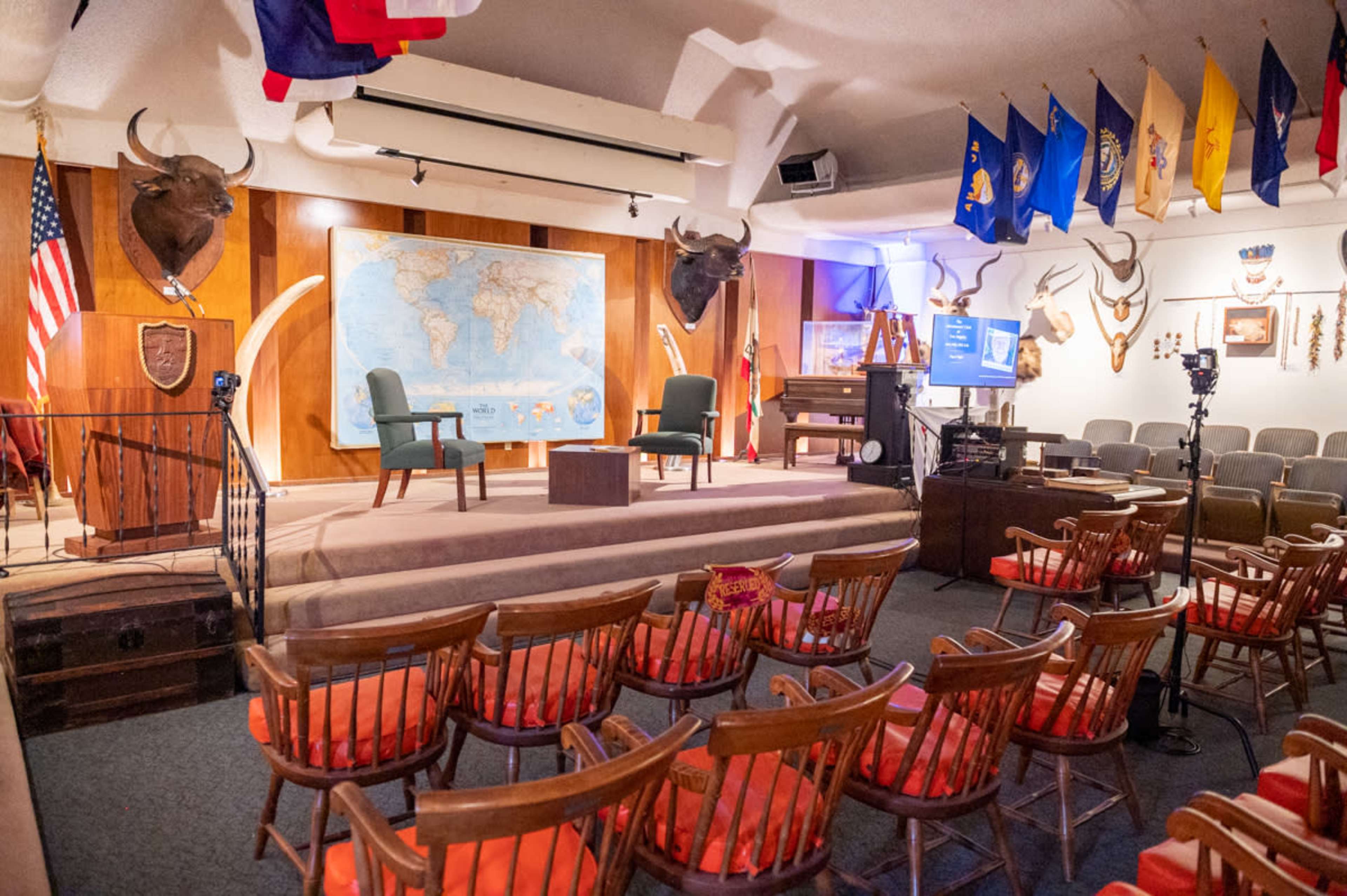 A stage setting with two chairs and a small table in front of a world map, surrounded by animal mounts and flags, with an arrangement of wooden chairs facing the stage.