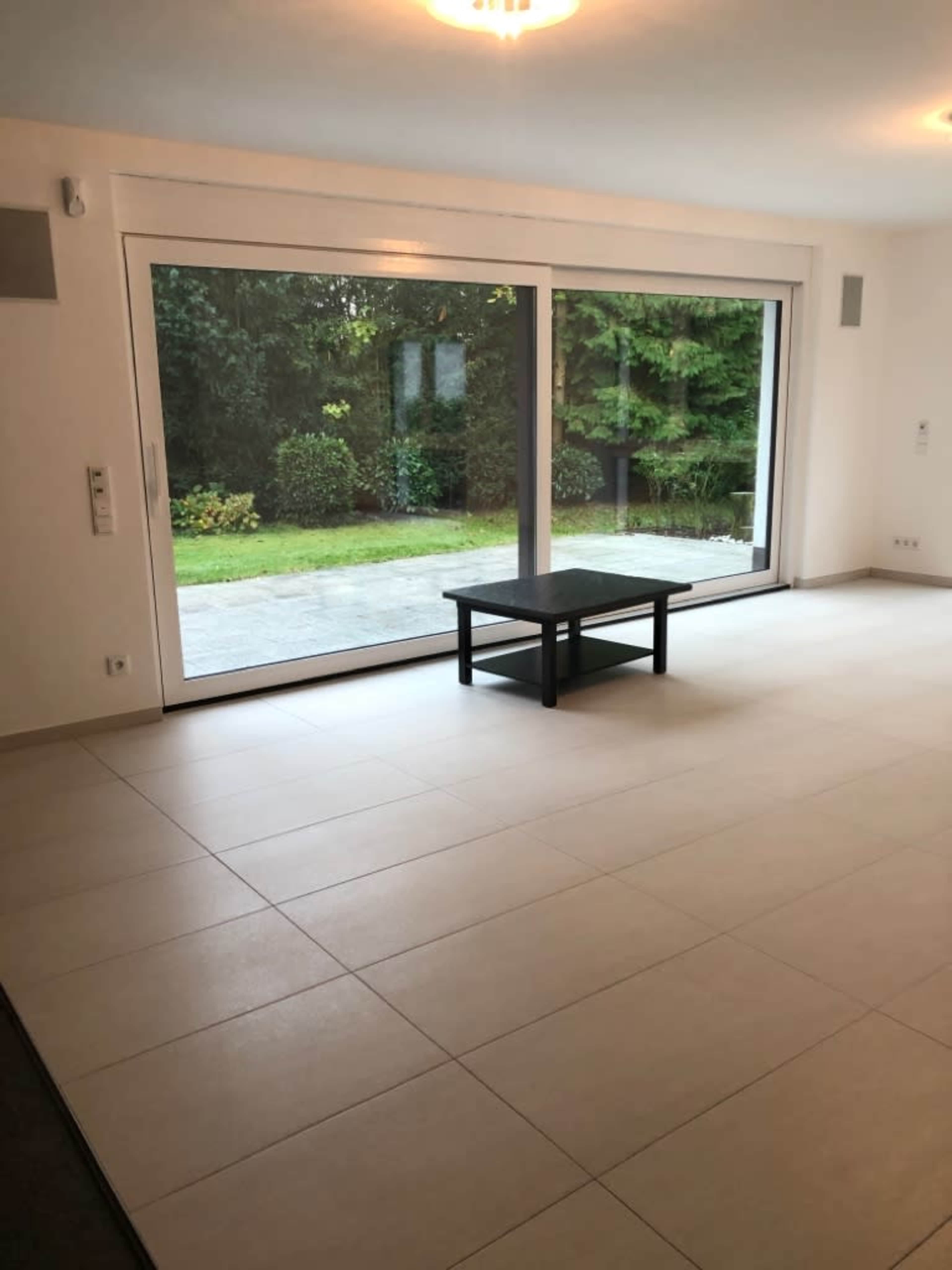 The image shows a spacious, empty room with a large sliding glass door opening to a garden area, featuring neutral-colored walls and tiled flooring.