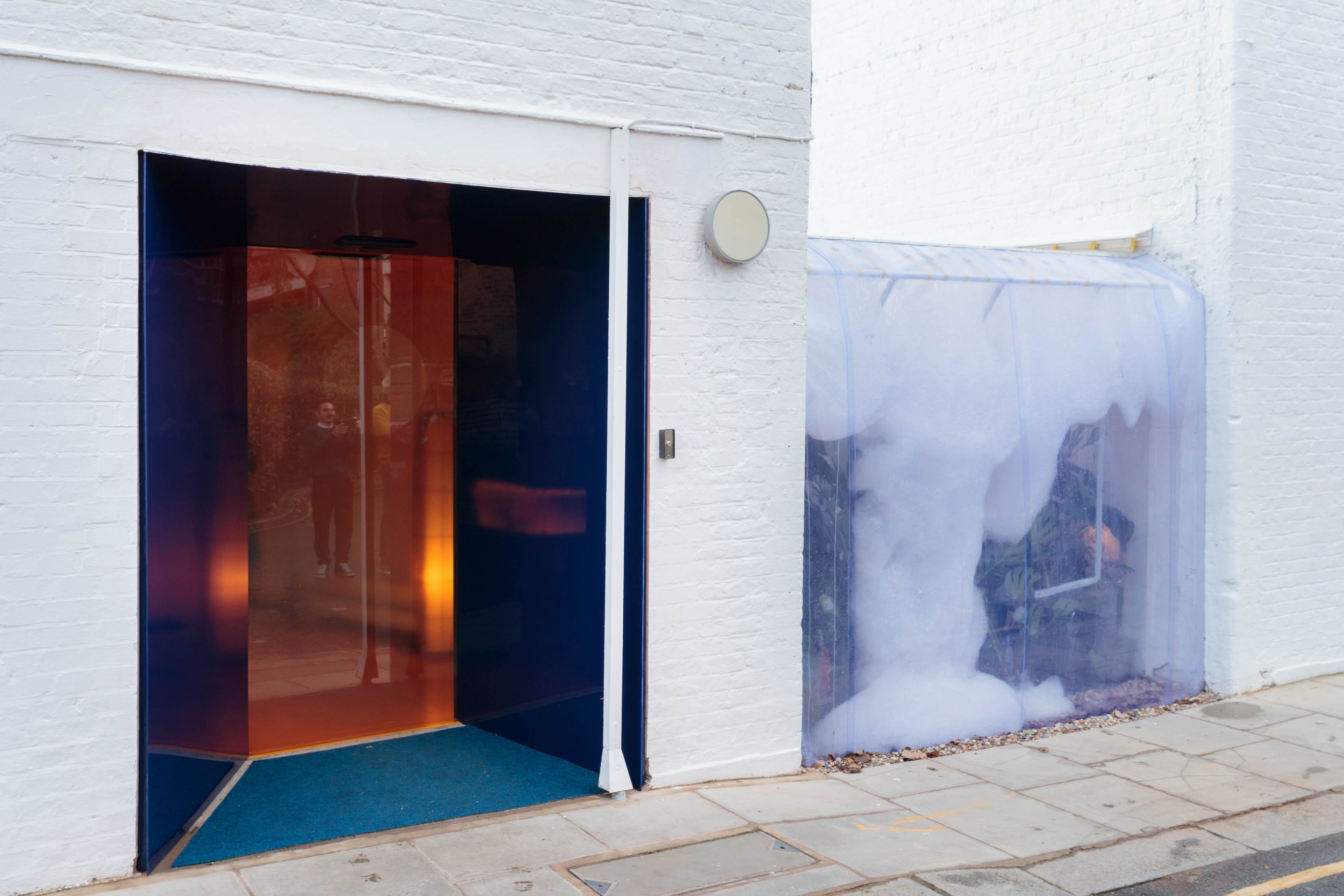 The image shows a modern entrance with a brightly colored glass door on the left, leading into a building, and a translucent, bubble-like structure on the right.