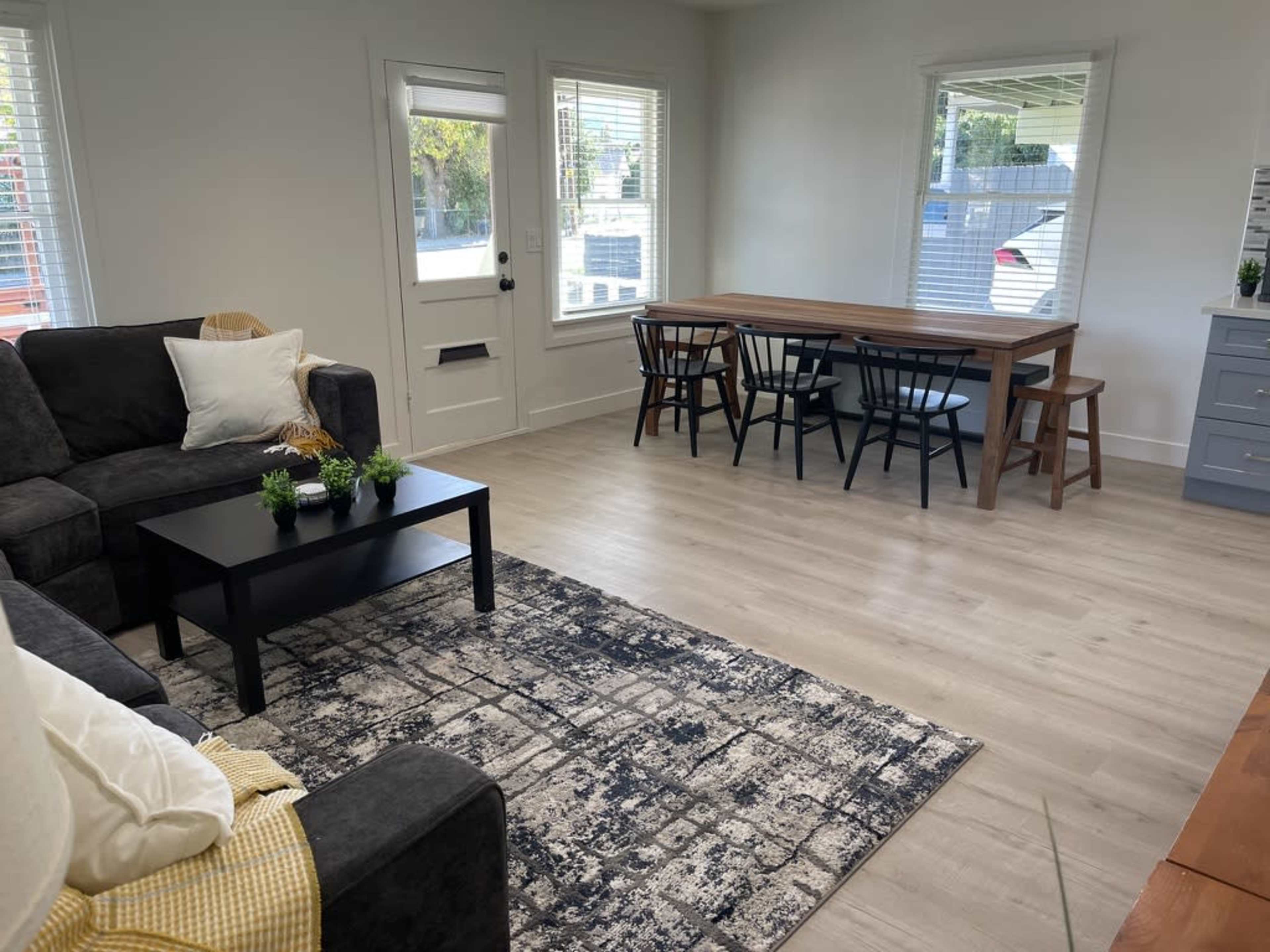 A spacious living room features a dark sofa, a coffee table, a dining table with chairs, and large windows letting in natural light.