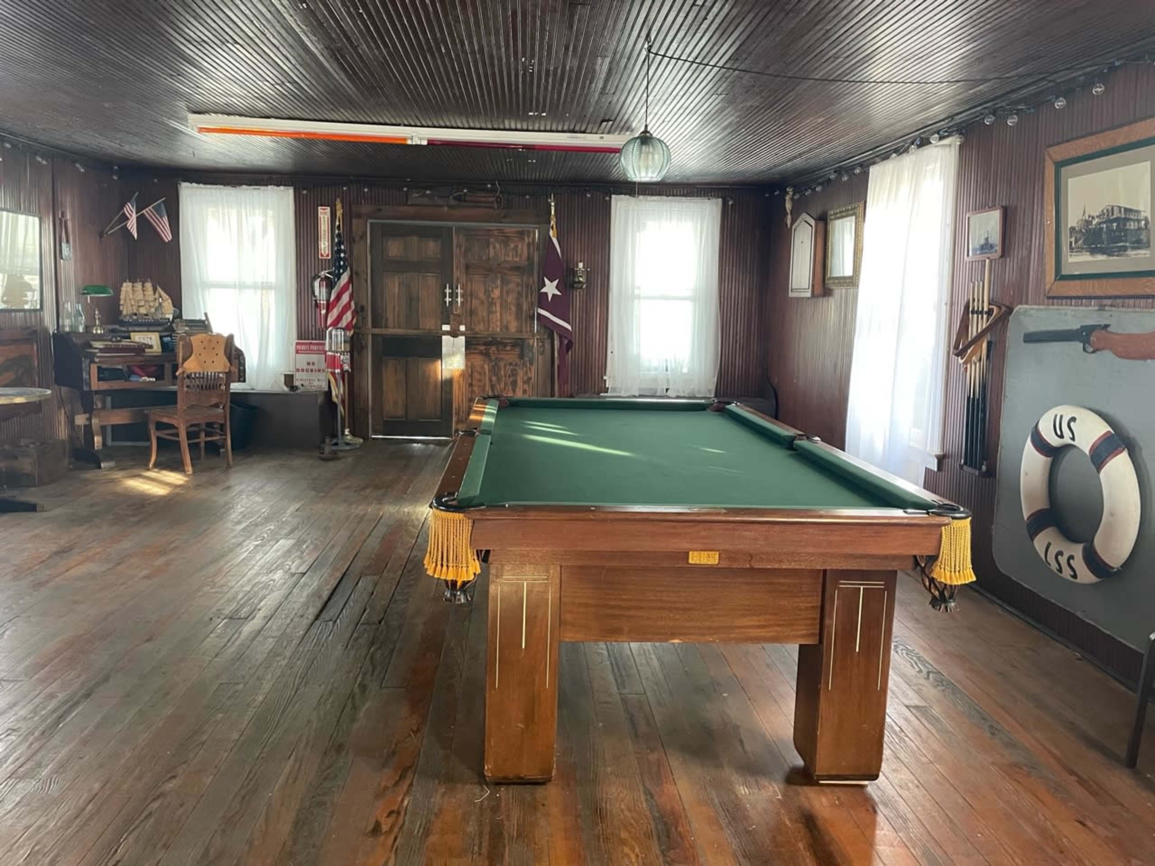 A room featuring a green felt pool table at the center, surrounded by wooden walls and traditional furnishings, with American flags and nautical decor.