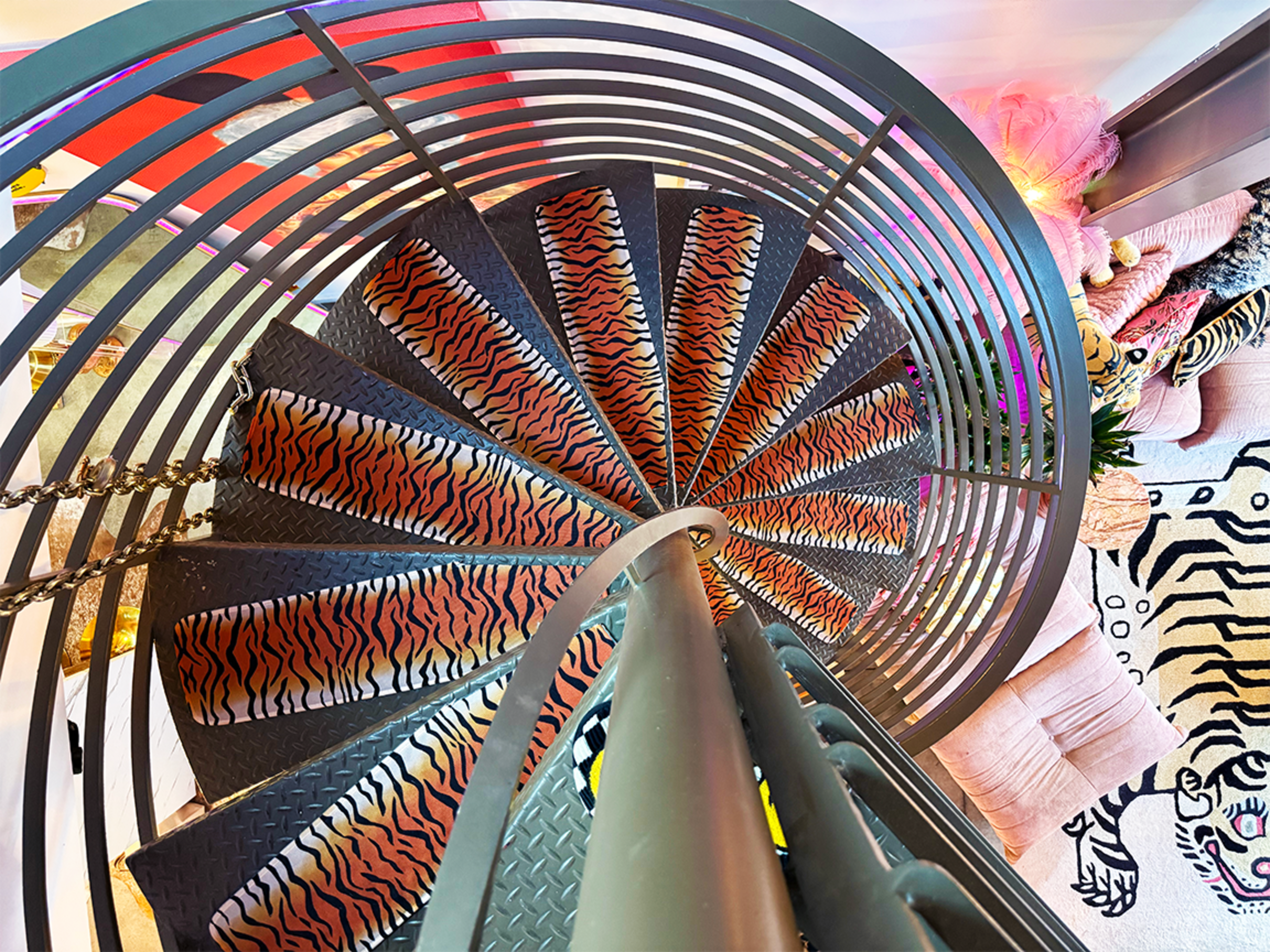 The image shows a spiral staircase with patterned treads and a handrail, leading down to a colorful room filled with various textiles and decorations.