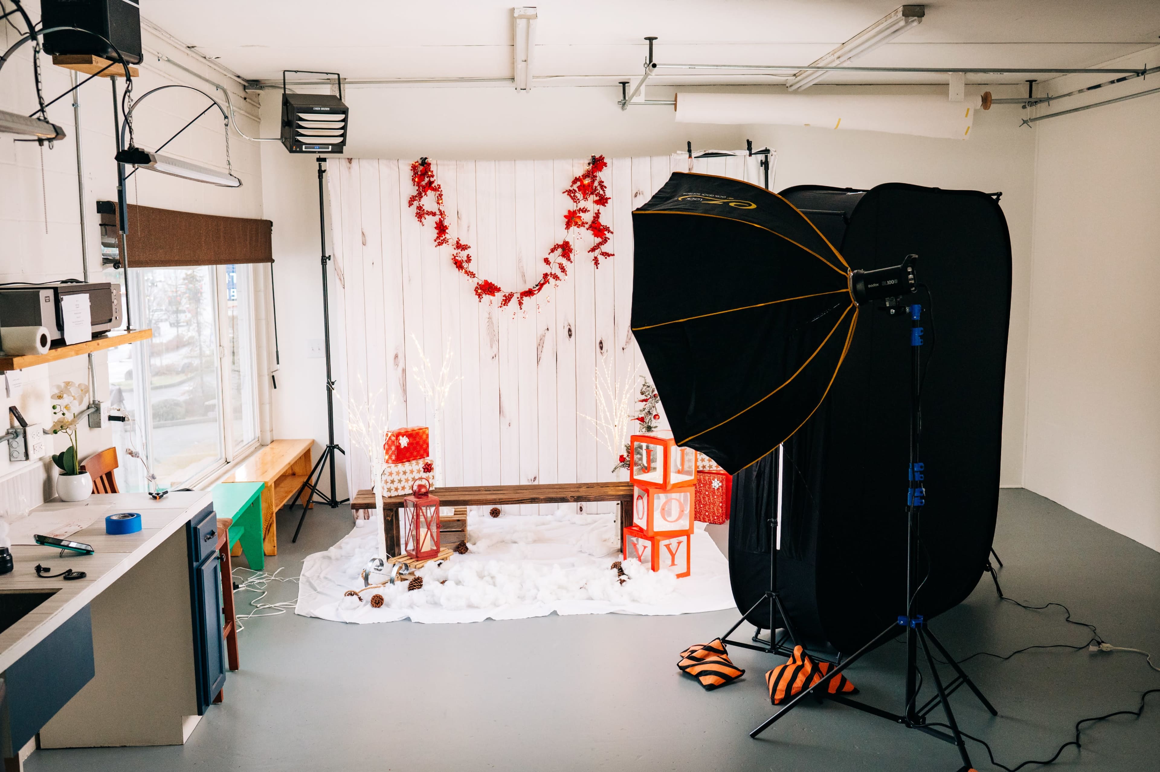 A photography studio is set up with a backdrop of white wooden panels adorned with red decorations, featuring a bench and Christmas-themed props.