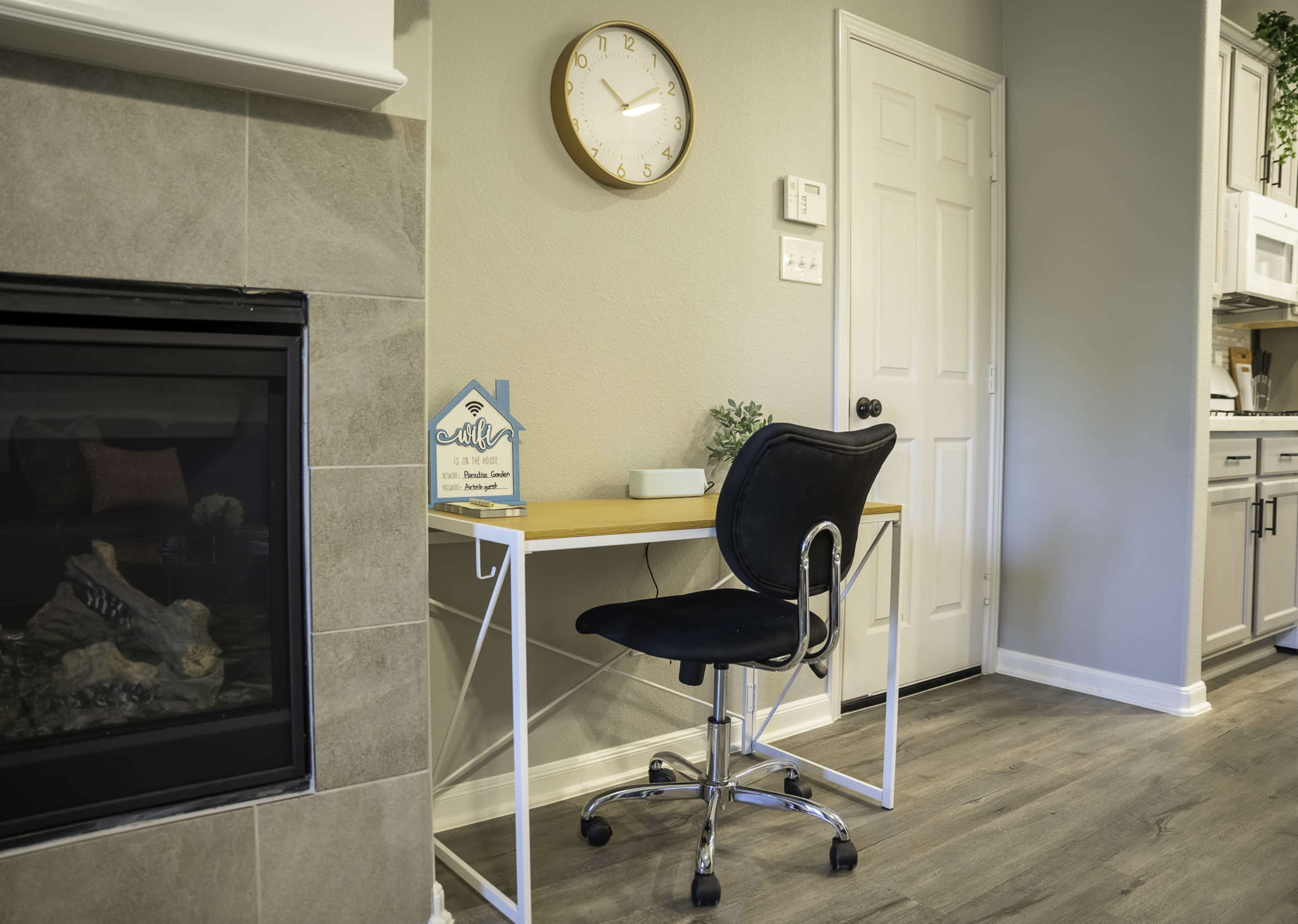 A modern home office space features a wooden desk with a black chair next to a fireplace and a wall clock.