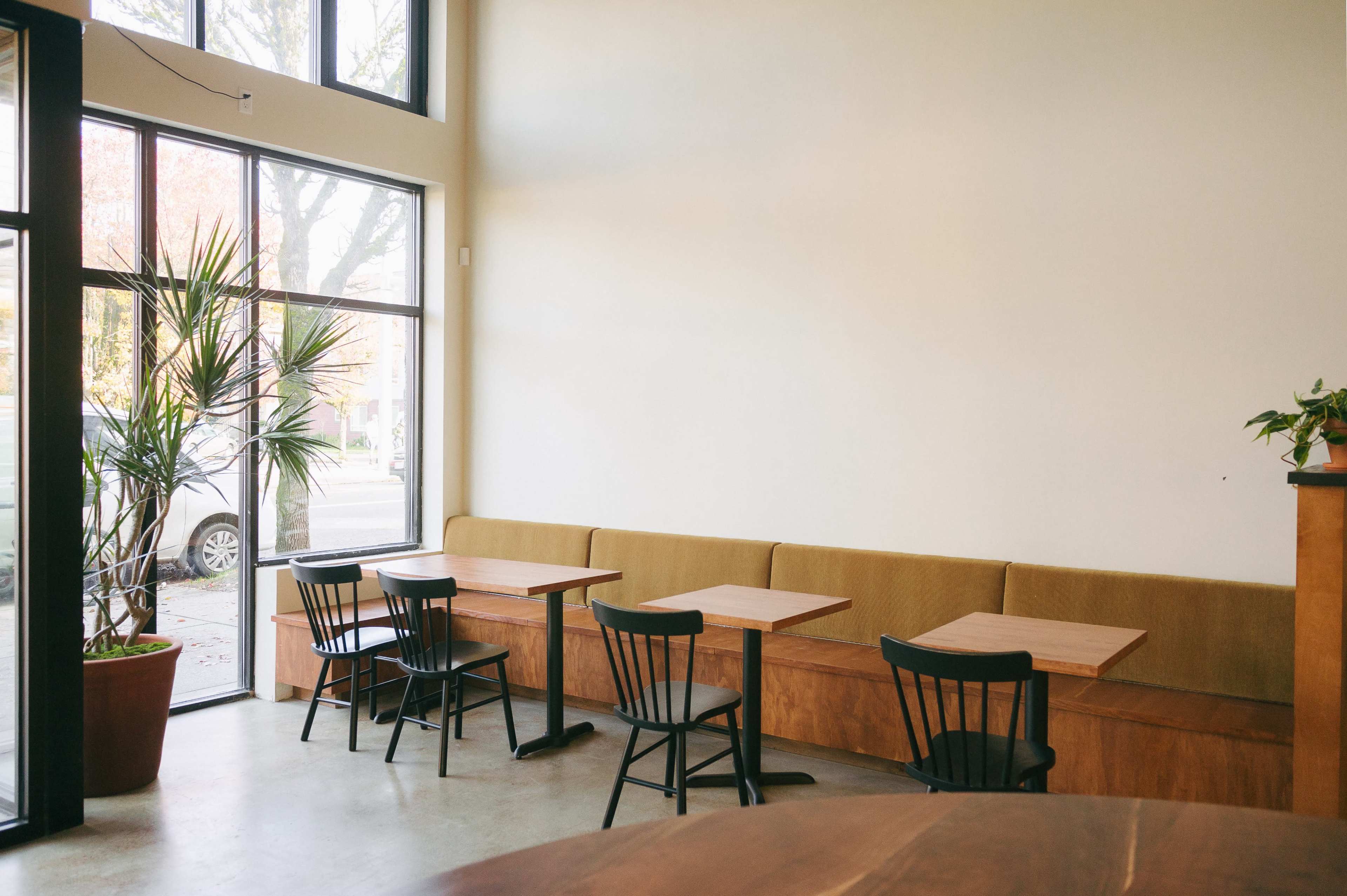 The image shows a minimalist café interior featuring a row of tables and chairs alongside a large window with a view of the street outside.
