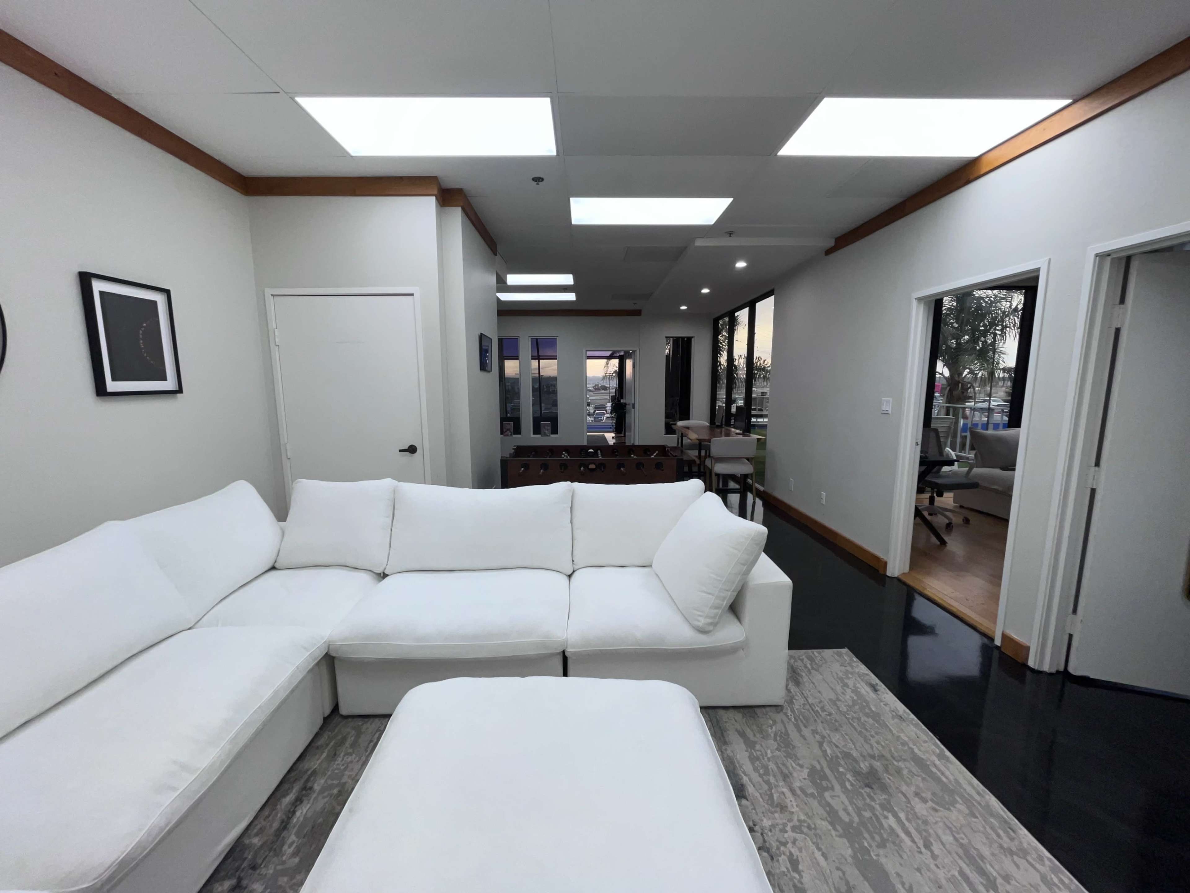 The image shows a modern, open-plan living space with a white sectional sofa and large windows revealing an outdoor view.